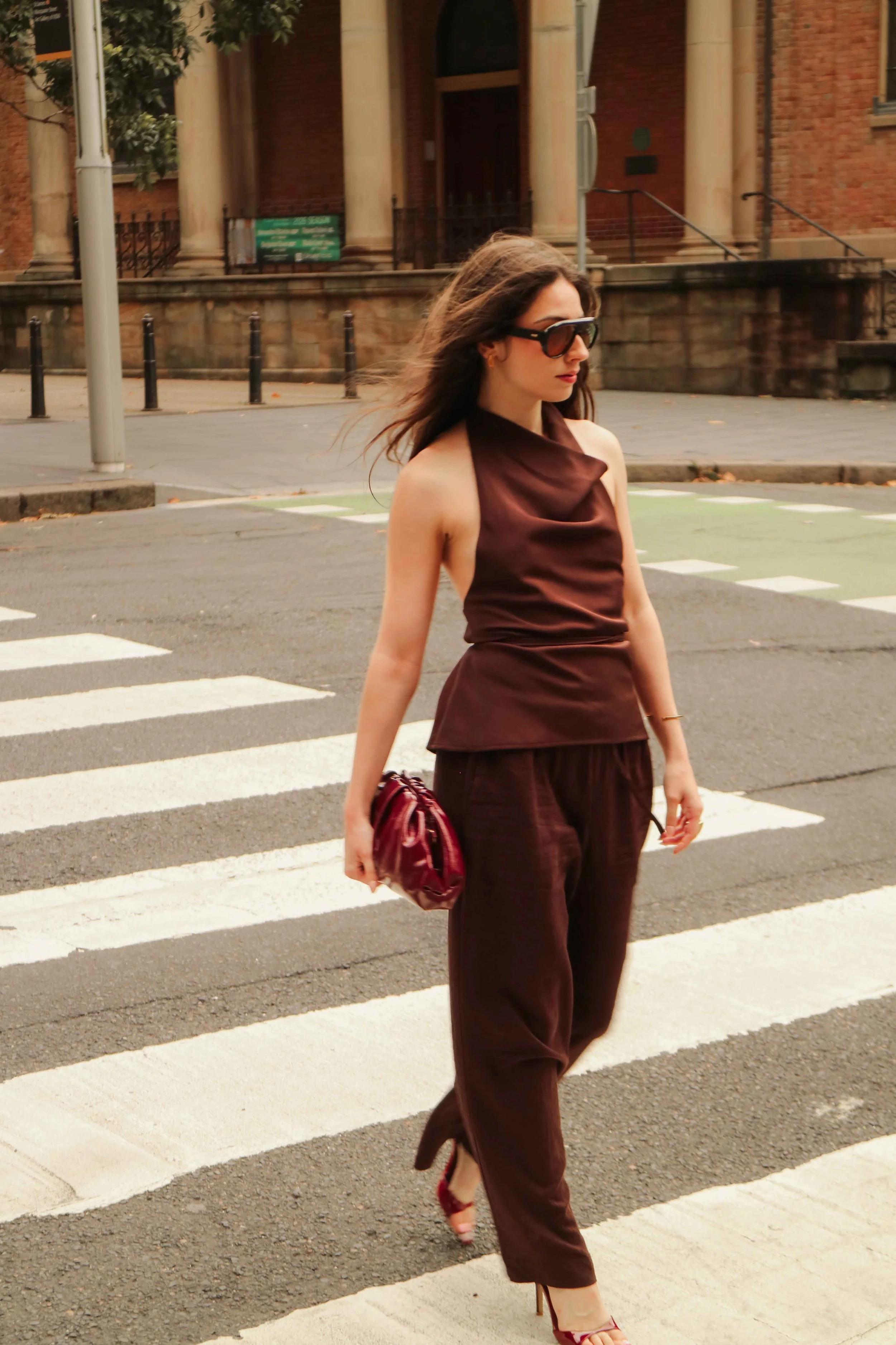 A woman crossing a street in an urban area, wearing a stylish sleeveless brown top and matching pants, holding a red clutch, with large sunglasses and high heels, with older brick buildings in the background.