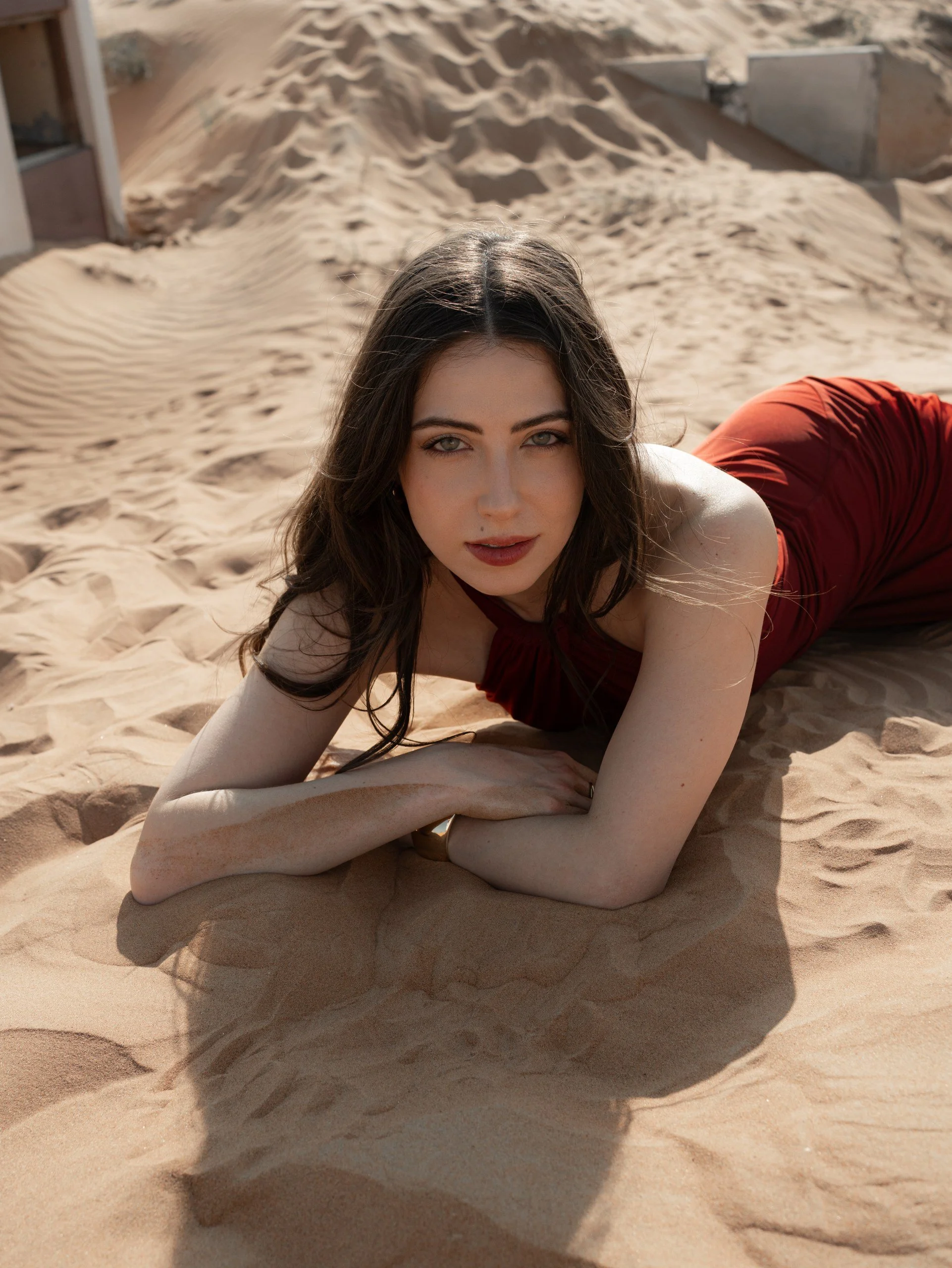 A woman in a red dress is lying on the sand in a desert, looking directly at the camera.