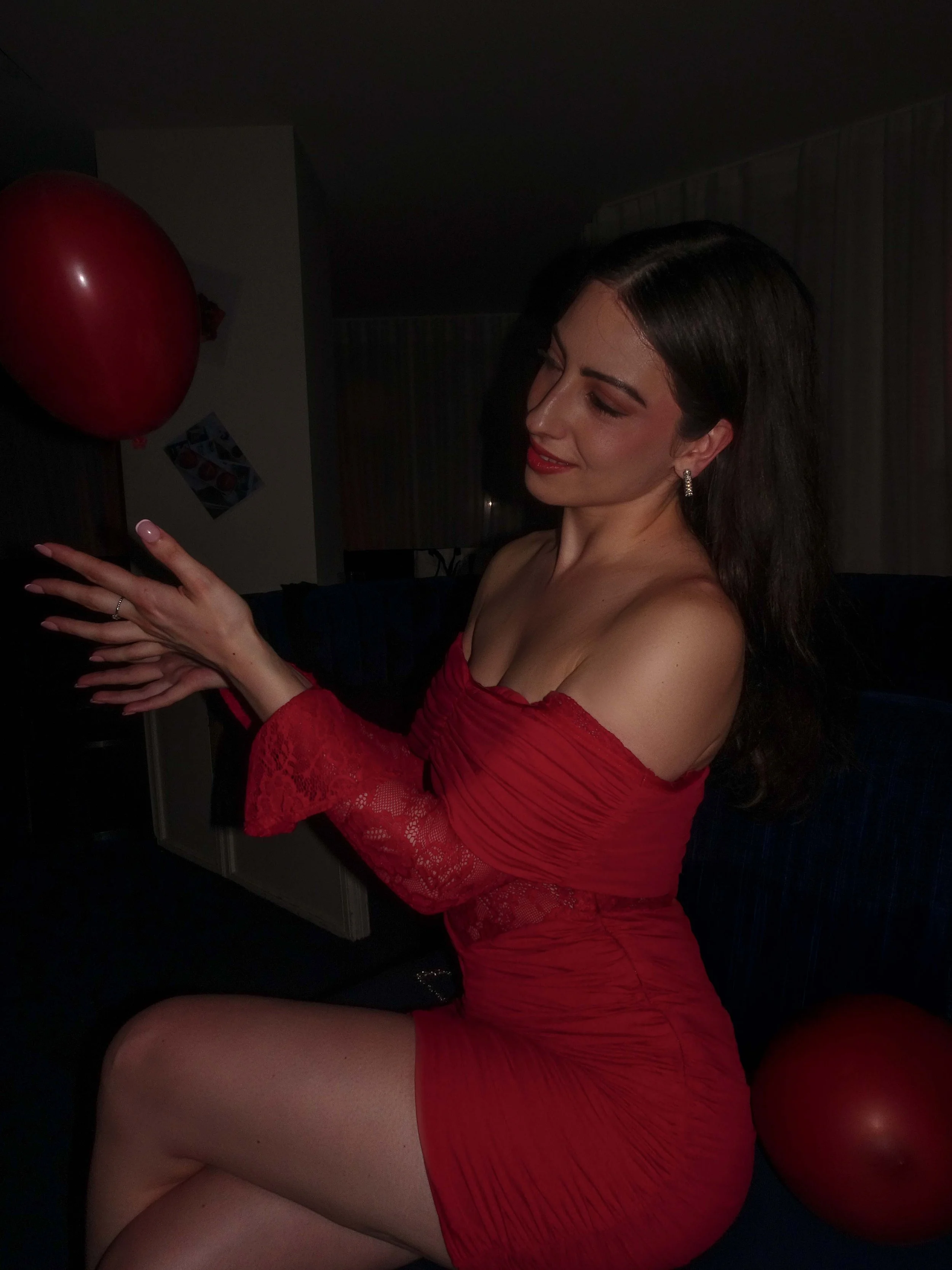 A woman in a red dress sitting indoors, holding a red balloon.