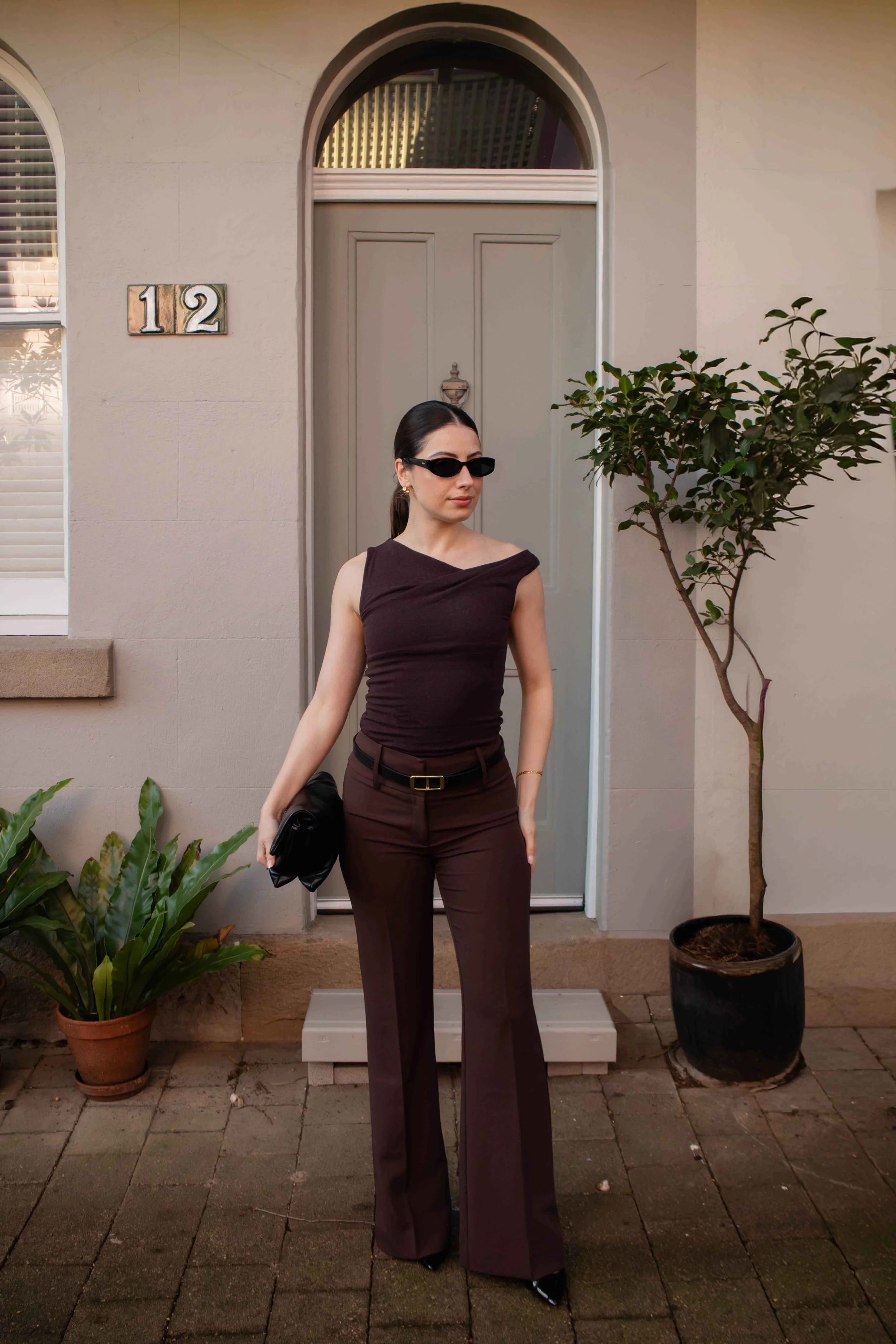 A woman wearing dark sunglasses, a black sleeveless top, and brown trousers stands in front of a light-colored house with the number 12 on it. She holds a black bag in her right hand and is positioned next to a potted plant with broad green leaves and a small tree in a black pot. The background includes a window with white blinds and concrete pavers on the ground.