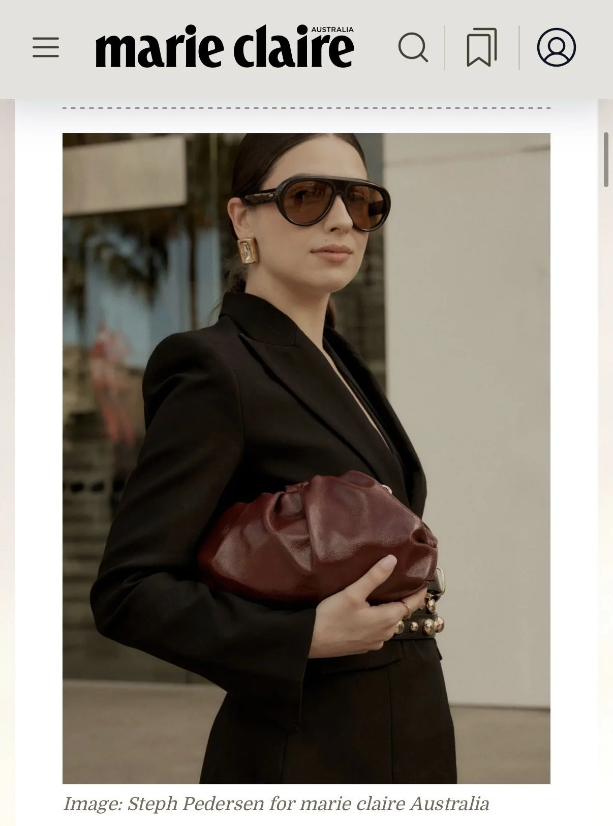 A woman with dark hair, wearing large sunglasses and gold earrings, clothes in dark colors, holding a red leather clutch bag, standing outdoors near a building.