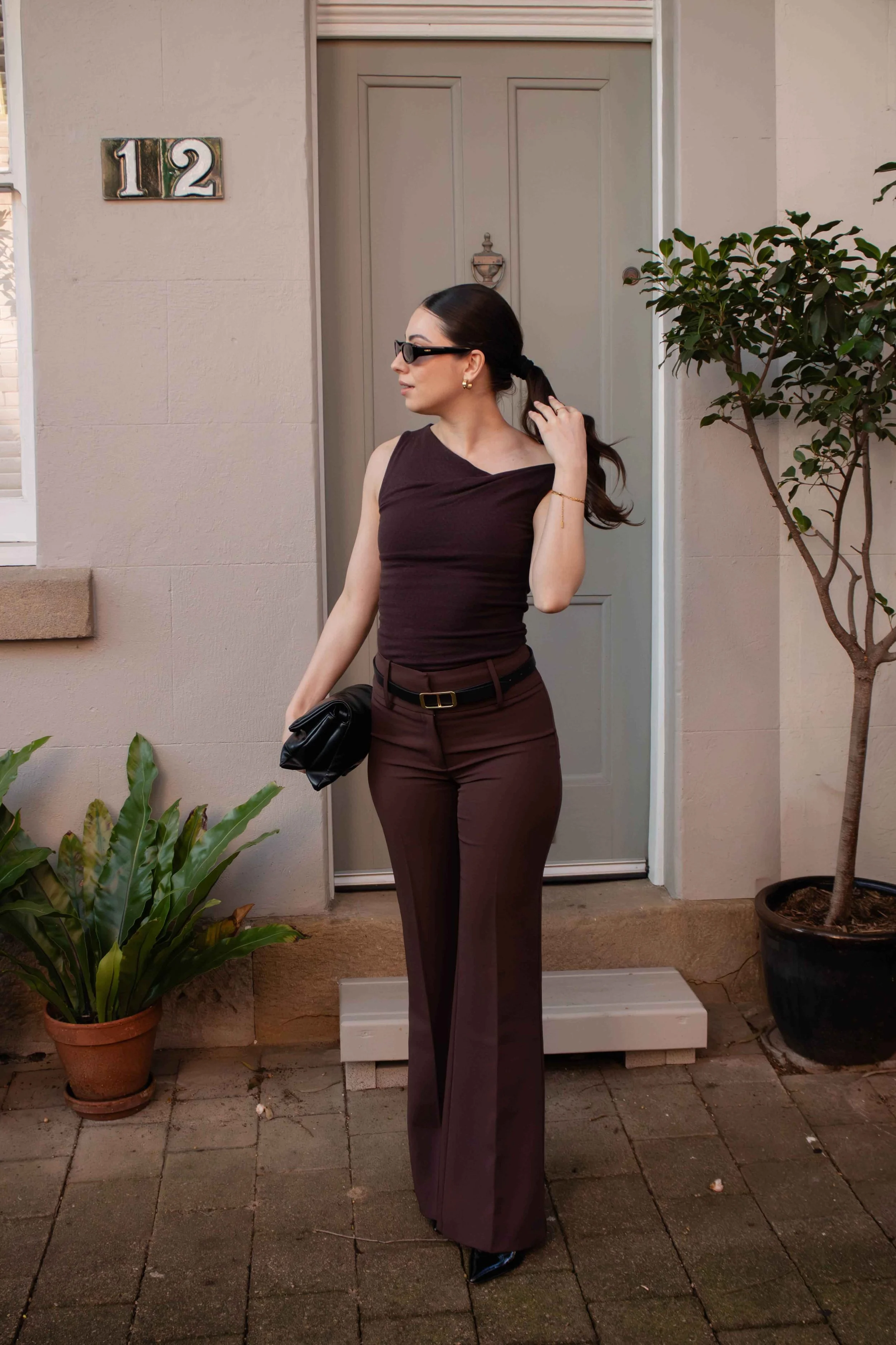 A woman with dark hair tied back, wearing sunglasses, gold earrings, a black sleeveless top, and brown high-waisted pants, stands outside on a brick patio near a beige wall with a house number "12" and a door with a knocker. She is holding a black purse in her left hand and adjusting her hair with her right hand, with a potted plant nearby.