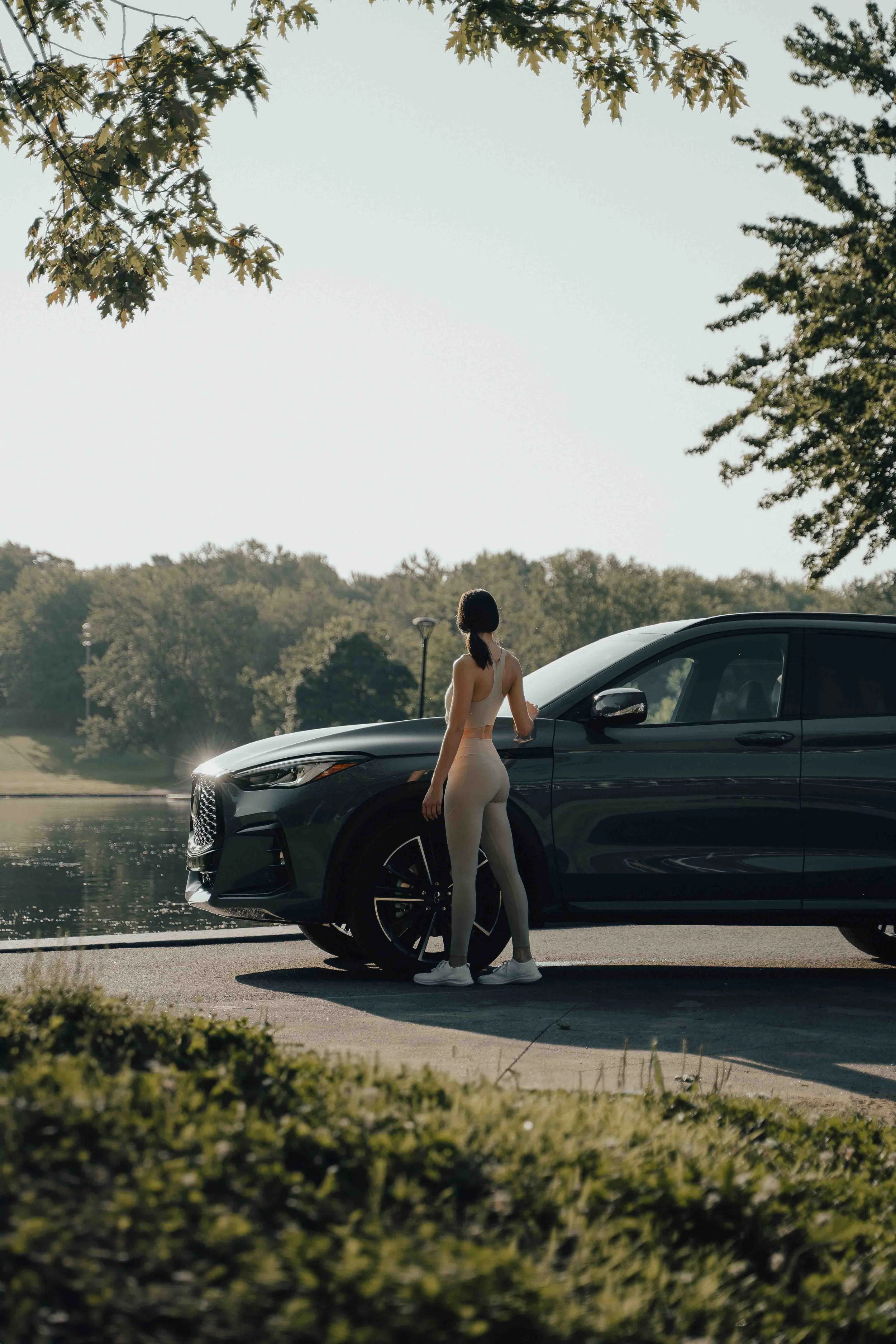 A woman in athletic clothing stands next to a black car by a lake, with trees and a clear sky in the background.