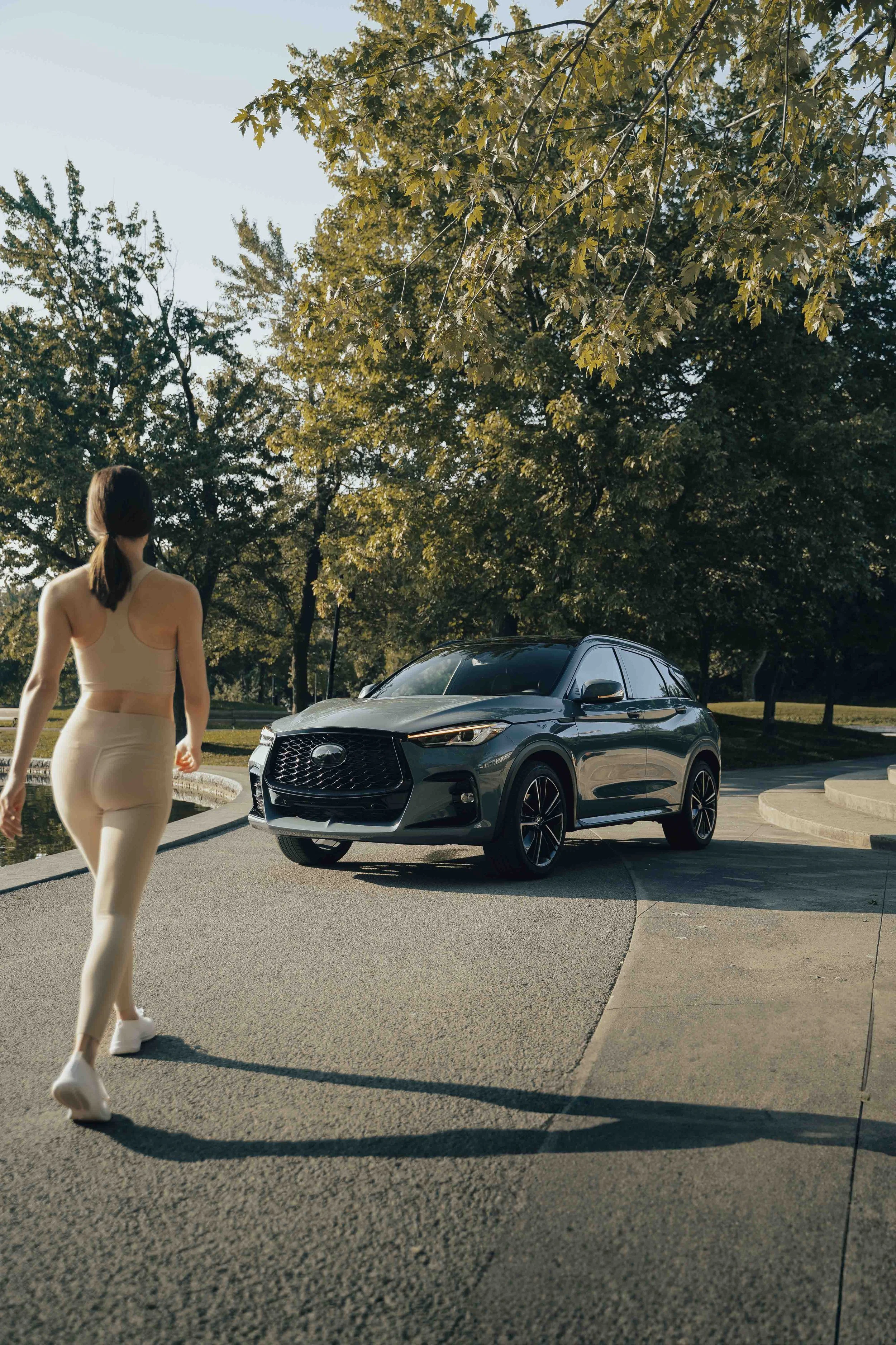 A woman walking near a parked gray SUV in a park-like area with trees and a fountain.