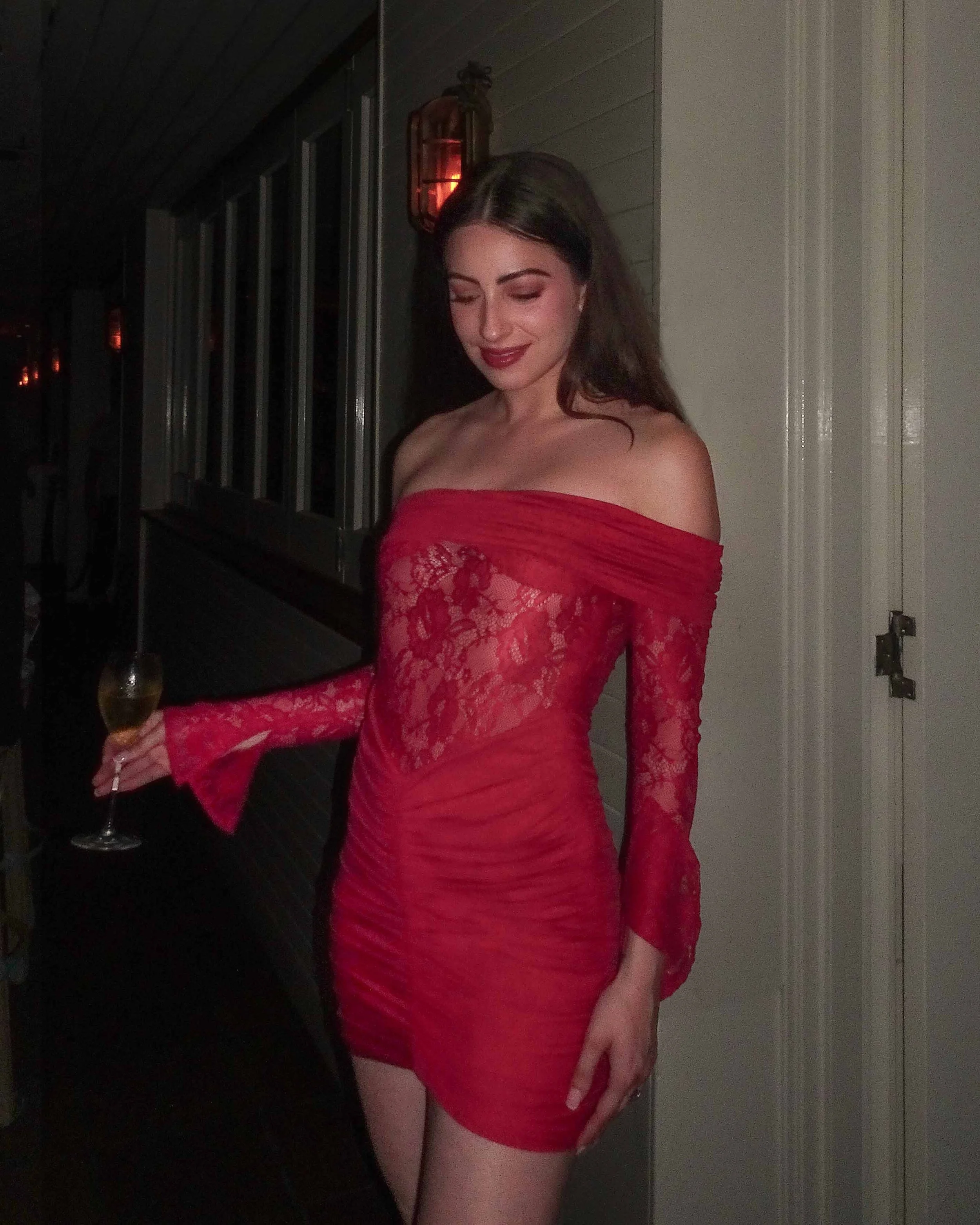 A woman in a red off-shoulder lace dress holding a glass of wine, standing in a dimly lit room with wall sconce lights.