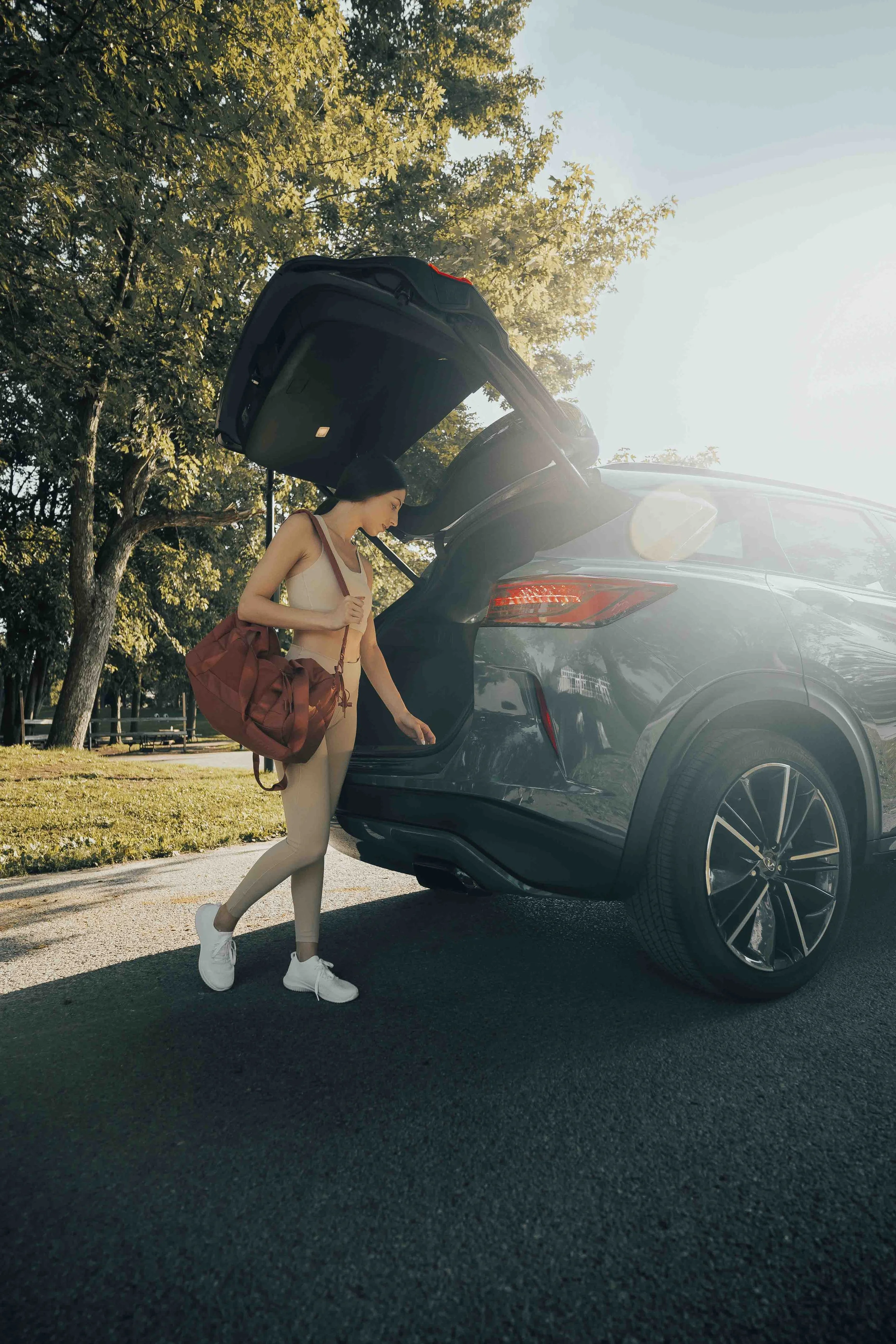 A woman loading a bag into the trunk of a black SUV parked on a sunny day in a park with trees.