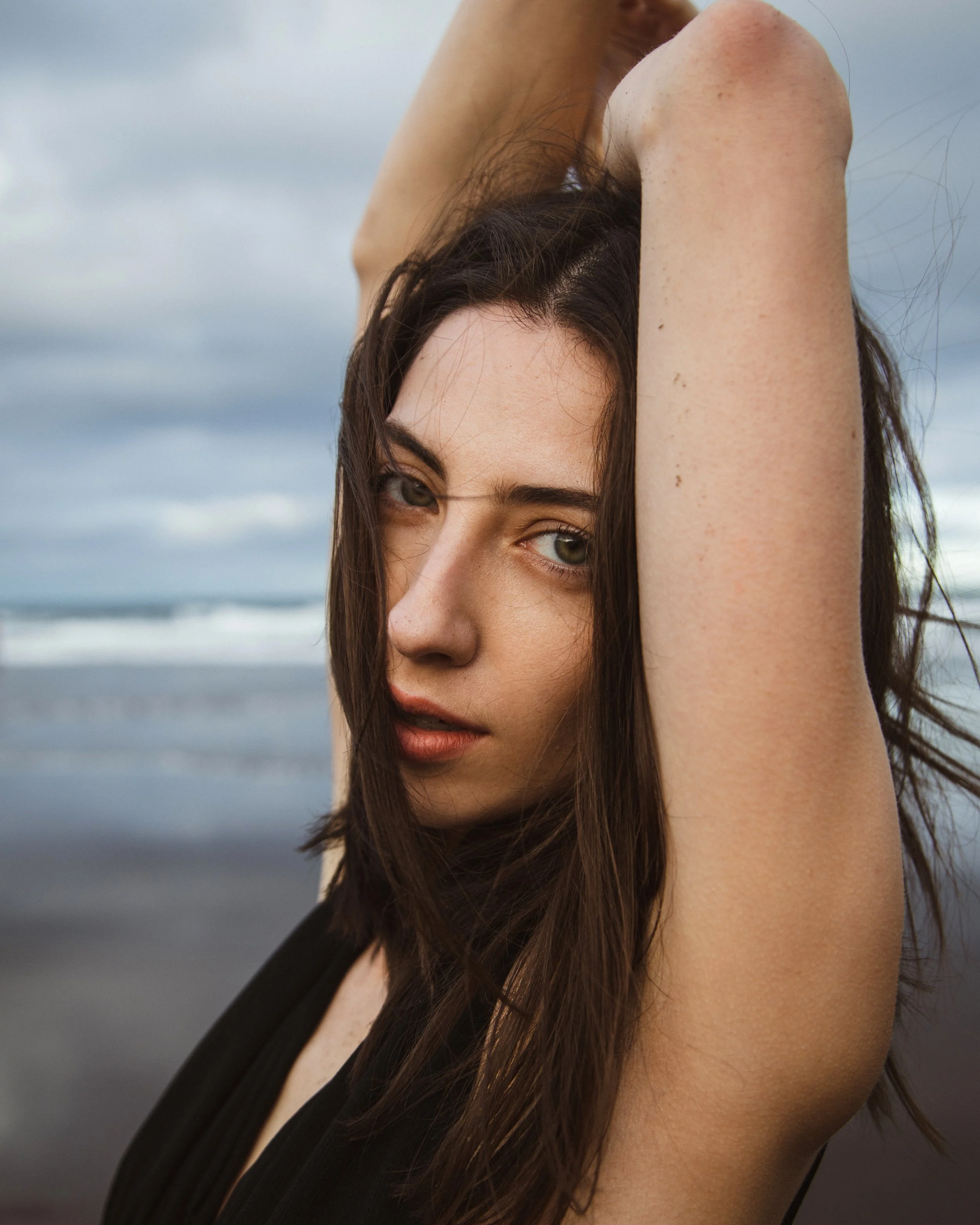 A woman with dark brown hair and green eyes posing outdoors at the beach with her arm raised above her head, looking into the camera.