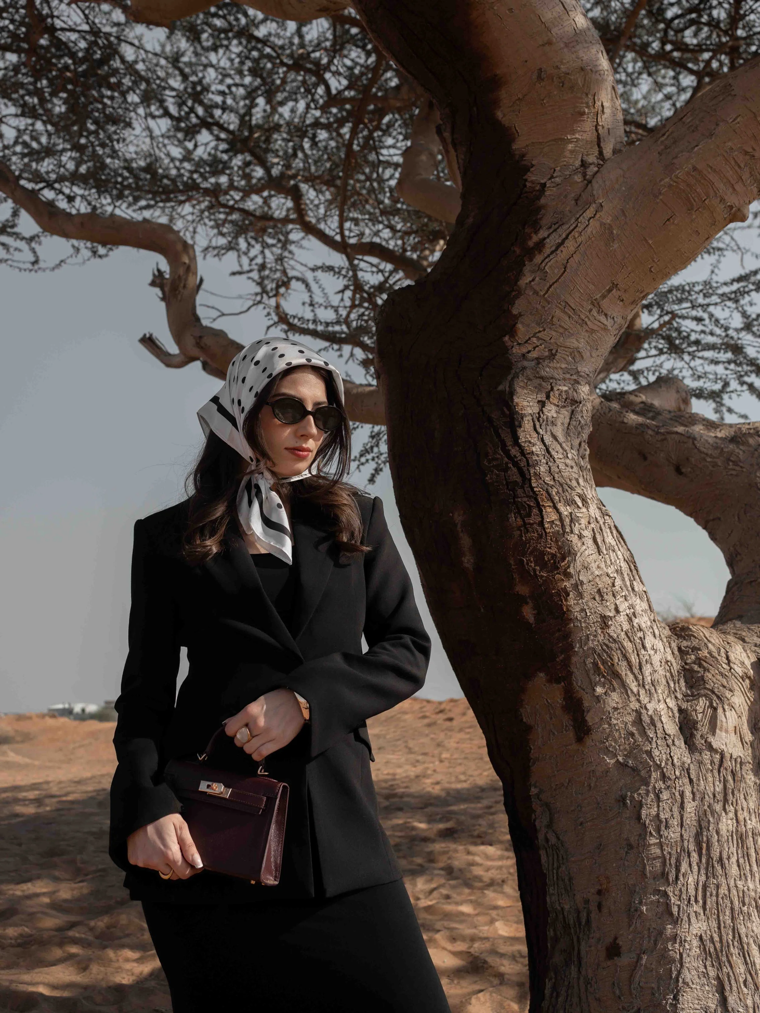 A woman in a black suit and polka dot headscarf standing beside a large tree in a desert landscape, wearing sunglasses and holding a dark handbag.