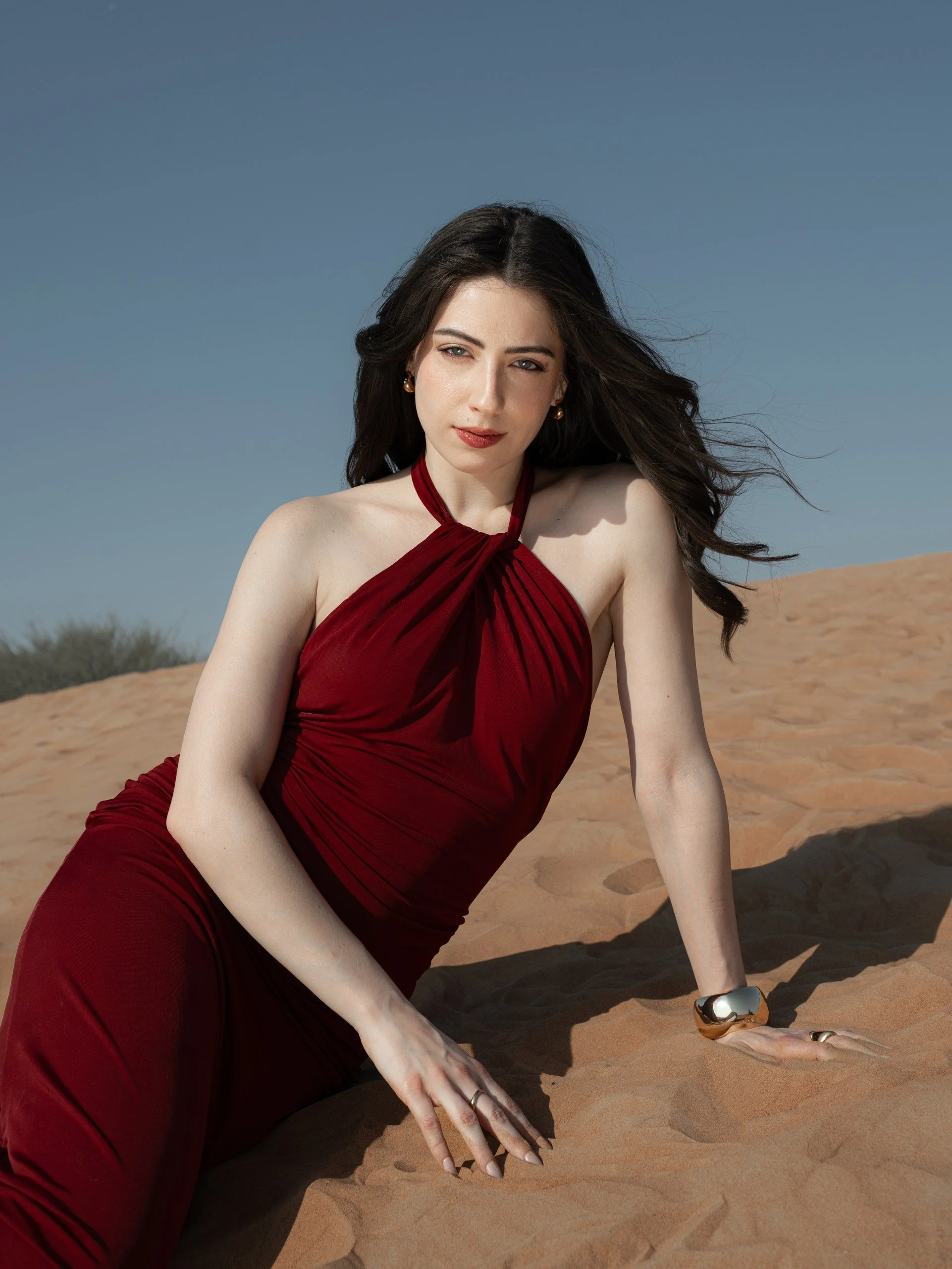 A woman in a red dress with halter neckline and pleats, sitting on sandy desert terrain, with wind-blown dark hair, posing under a clear blue sky.