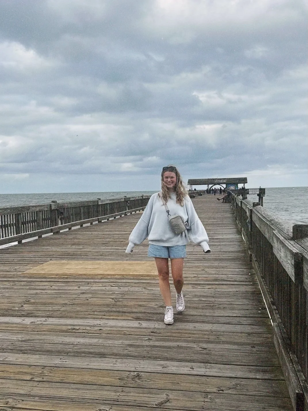A person walking on a wooden pier over the ocean, wearing a light-colored sweater and denim shorts, with cloudy skies in the background.