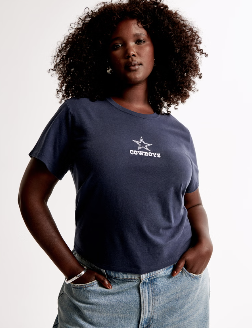 Person with curly hair wearing a navy t-shirt with 'Cowboys' and a star logo, paired with light blue jeans.