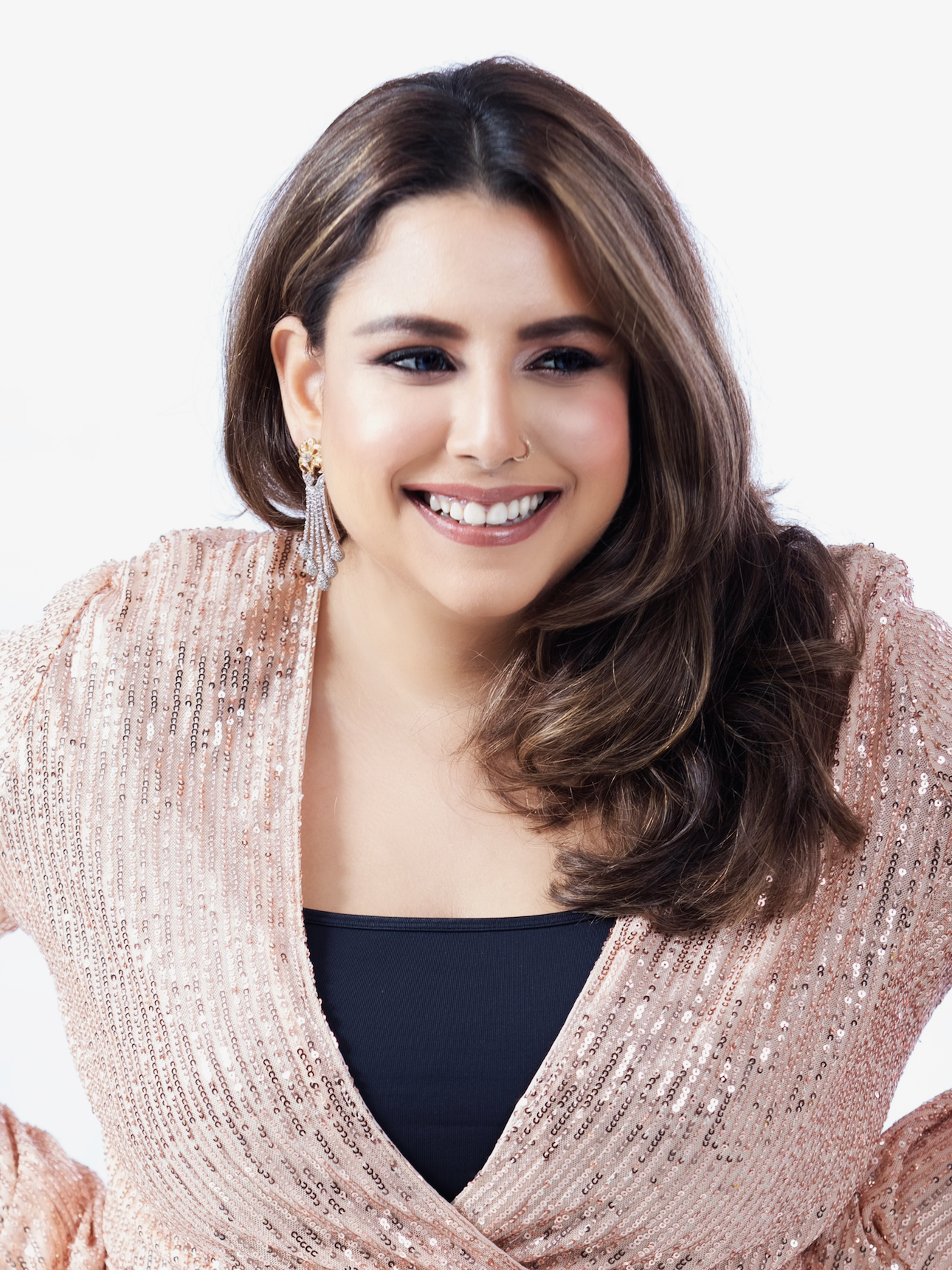 A woman with brown hair styled in loose waves, wearing a sparkly pink blazer over a black top, with earrings and a nose ring, smiling against a white background.