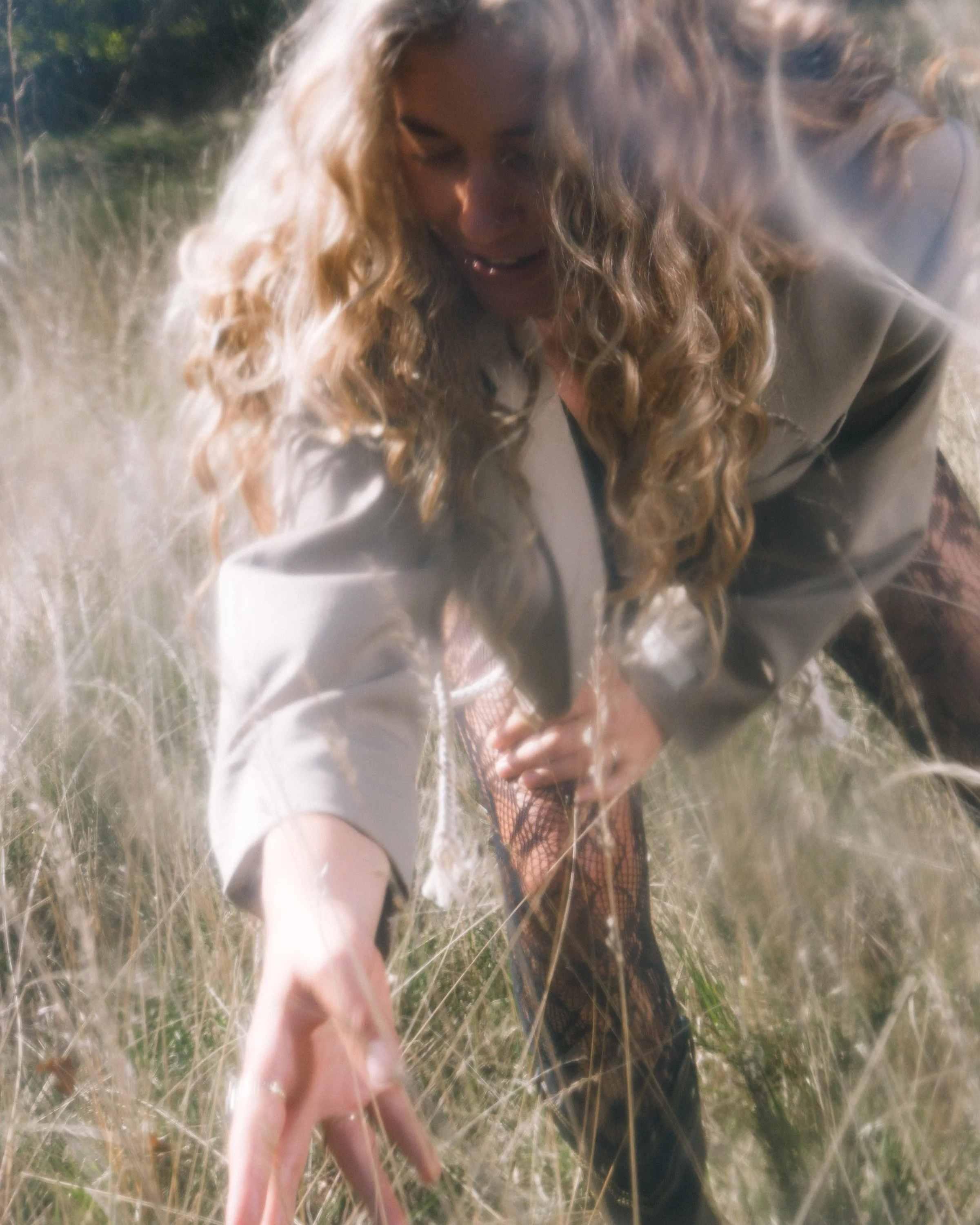 Vrouw met lang, krullend blond haar buigt door gras en strekt haar hand uit naar de camera. Ze draagt een beige jas en visnetkousen.