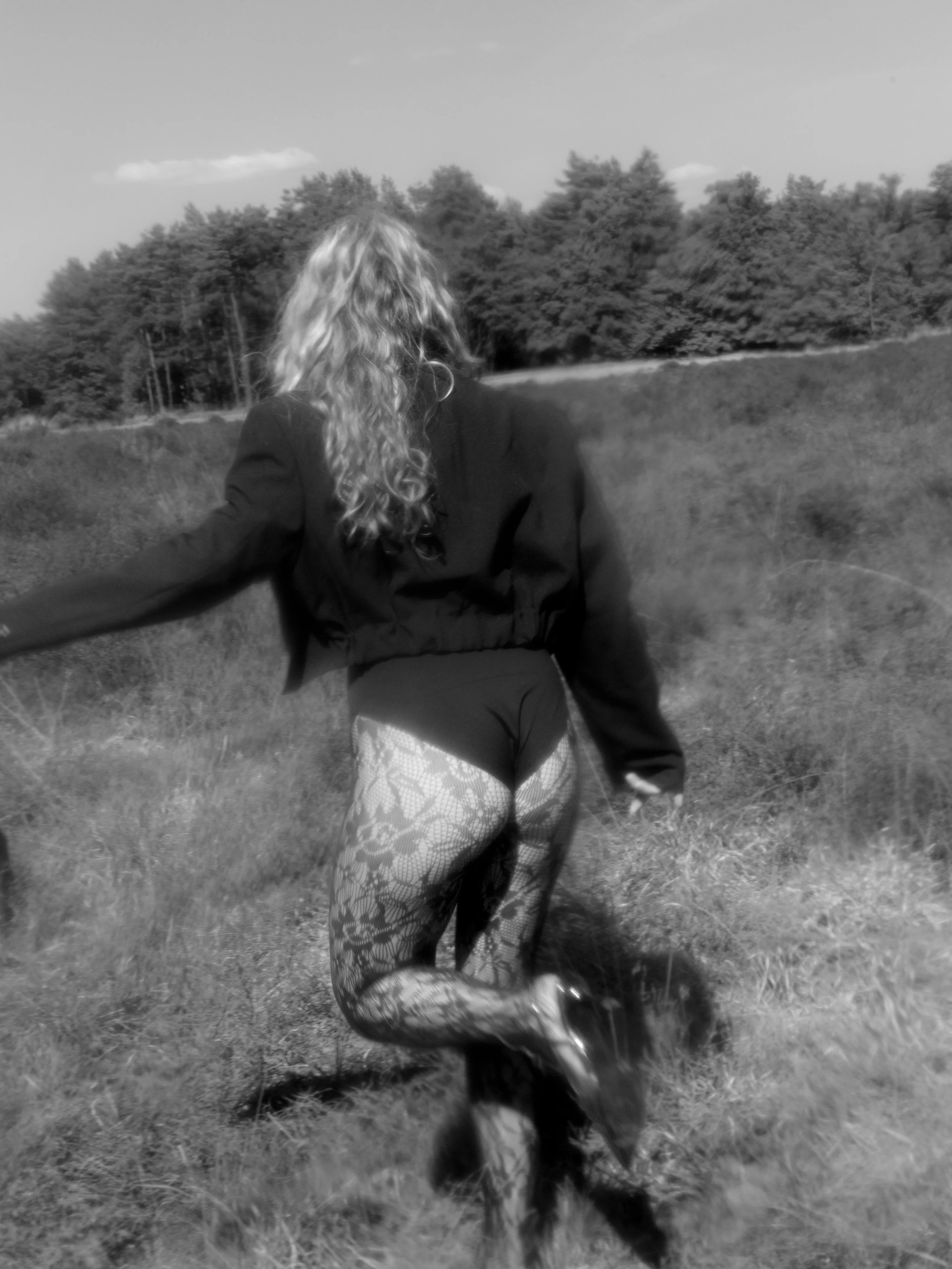 Een persoon met lang, golvend haar die in een veld staat, gekleed in een blazer, korte broek en opvallende panty's.
