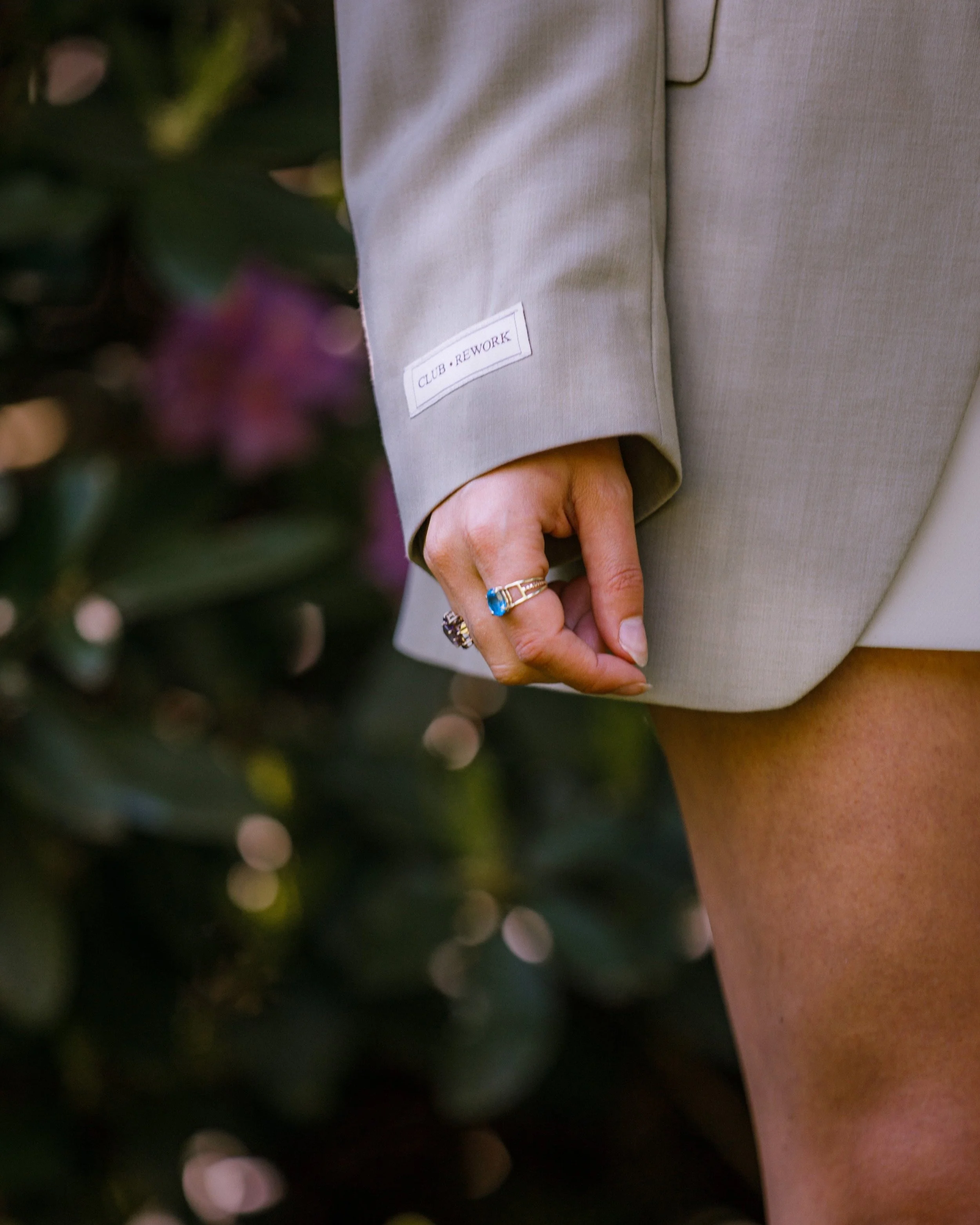 Delen van een persoon in een beige blazer met een label op de mouw die zegt 'CLUB · REWORK', een hand met sieraden, en een blote been. Op de achtergrond groene bladeren en bloemen.