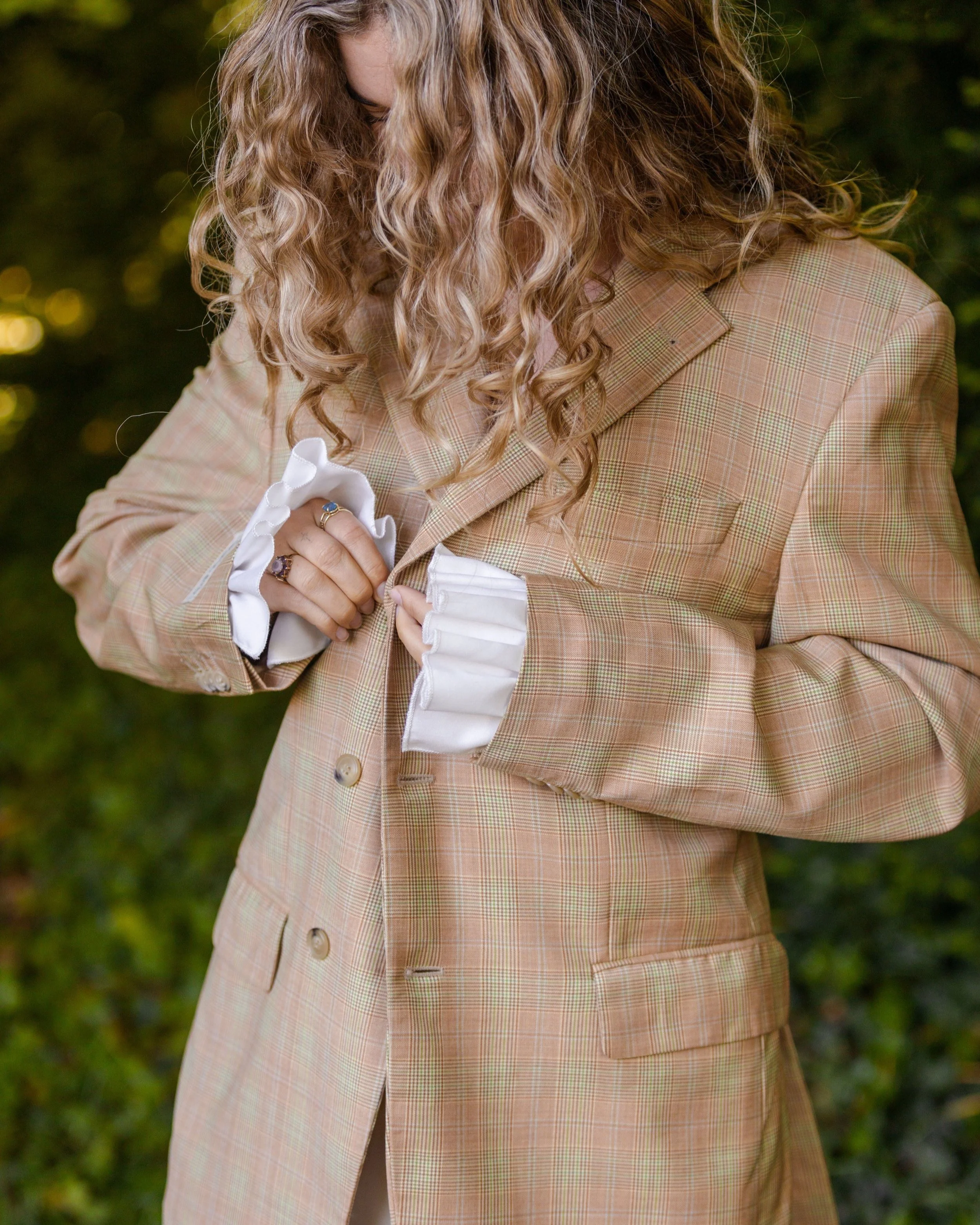 Dame met lang, krullend haar die een geruit, beige blazer draagt terwijl ze knoopt aan haar jas. Ze heeft een ring aan haar vinger en witte, gerufflede blousen met lange mouwen onder haar blazer.