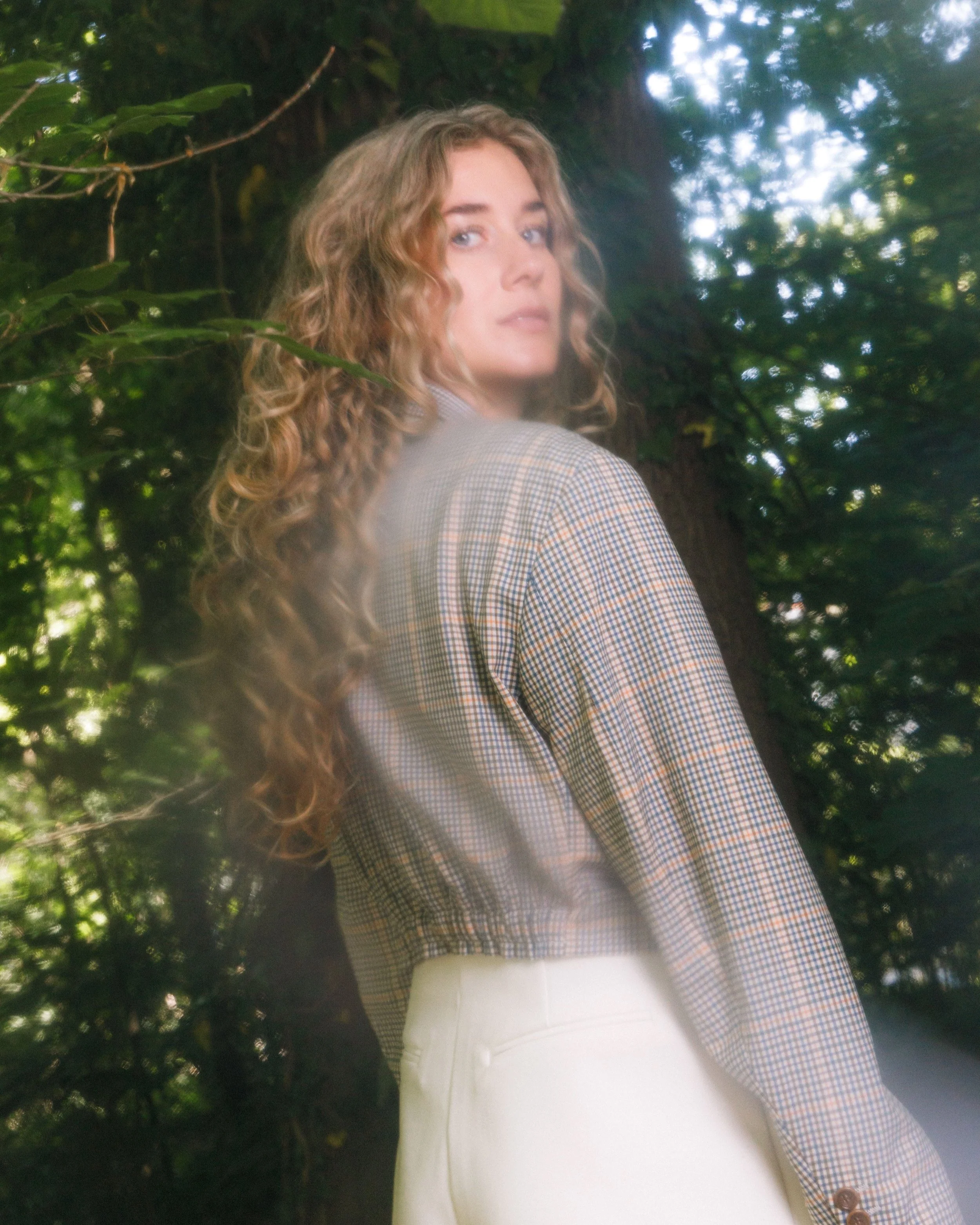 Een vrouw met lange, krullende haar kijkt over haar schouder naar de camera in een bosrijke omgeving.