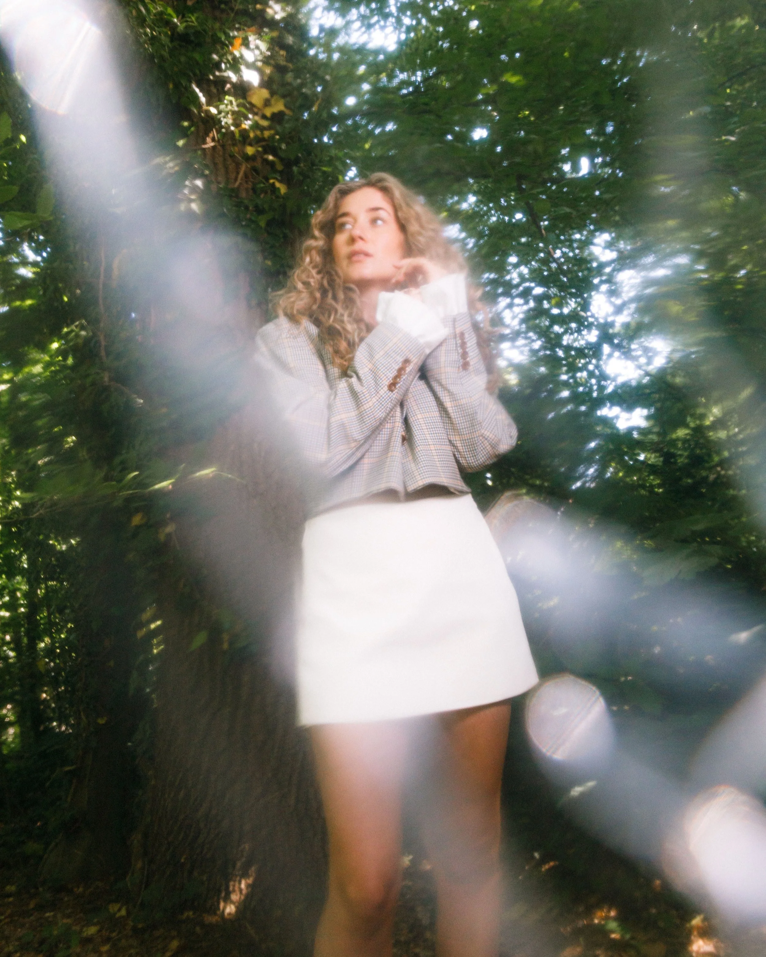 Een vrouw met gekruld haar staat in een bos, getooid in een grijs geruite blazer en een witte rok, omgeven door groene bladeren.