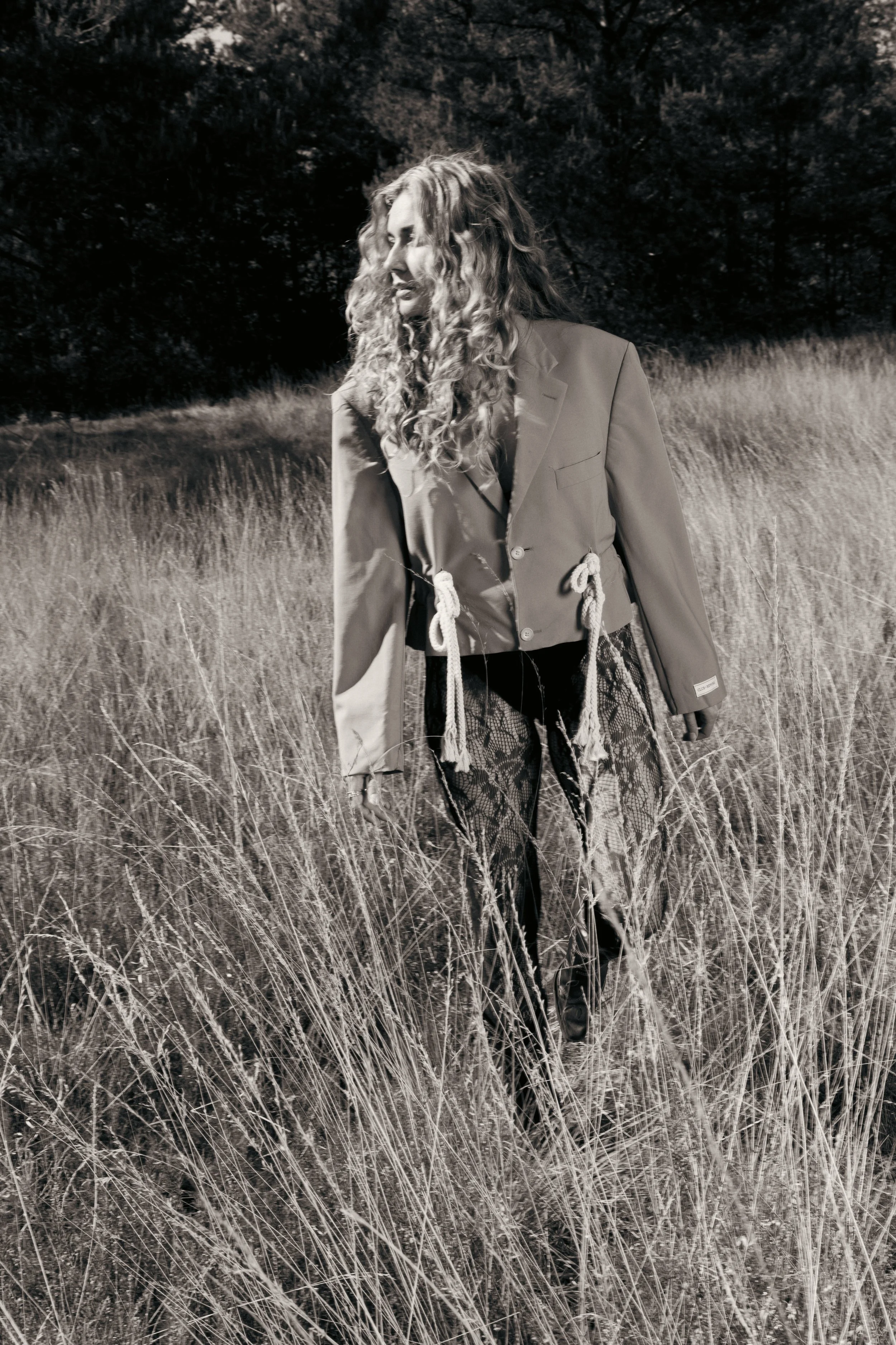 Een vrouw met lang, golvend haar in een veld met hoog gras, gekleed in een blazer en opvallende, doorzichtige broek met een print.