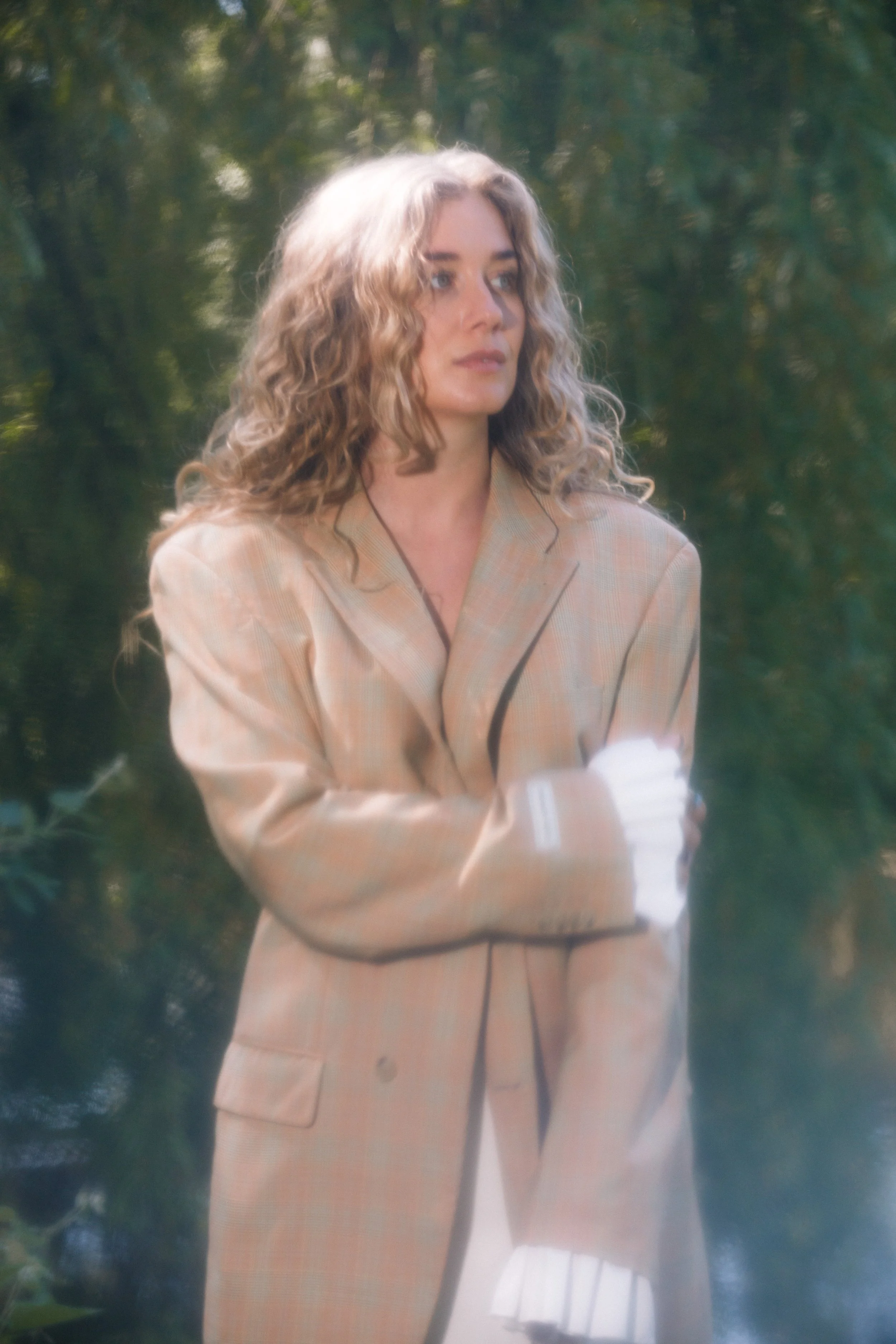 Een vrouw met golvend blond haar draagt een beige geruit blazer en witte handschoenen, staat buiten in een groene, bosachtige omgeving.