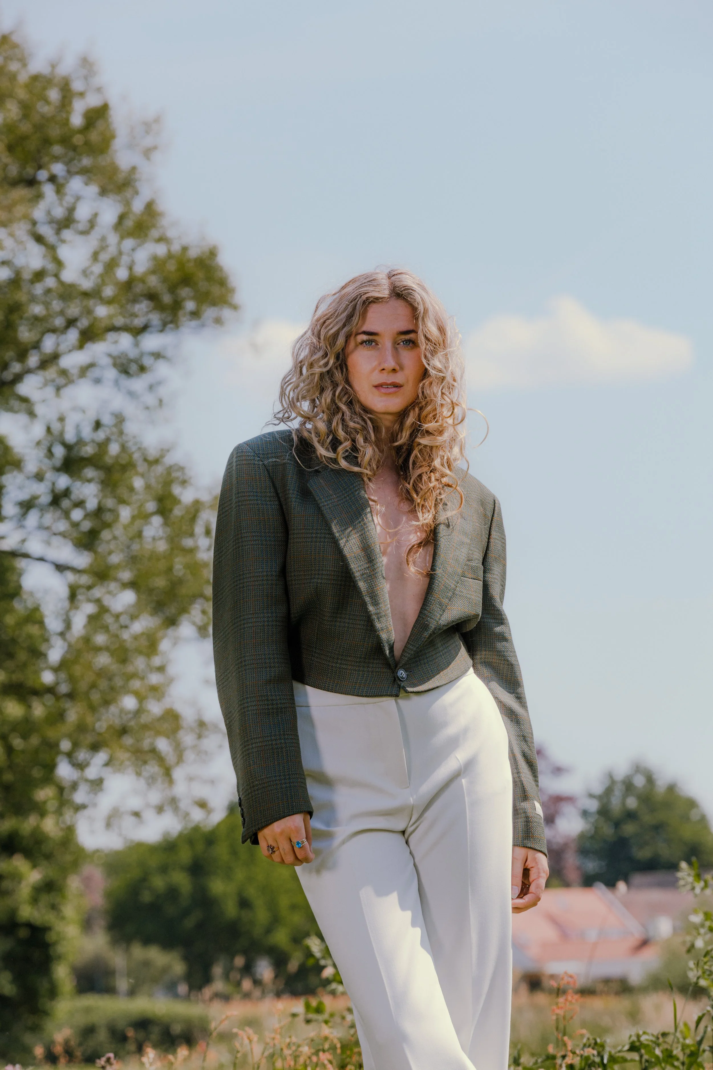 Vrouw met lang, krullend blond haar draagt een groene blazer en witte broek, staat in een open veld met bomen en huizen op de achtergrond.