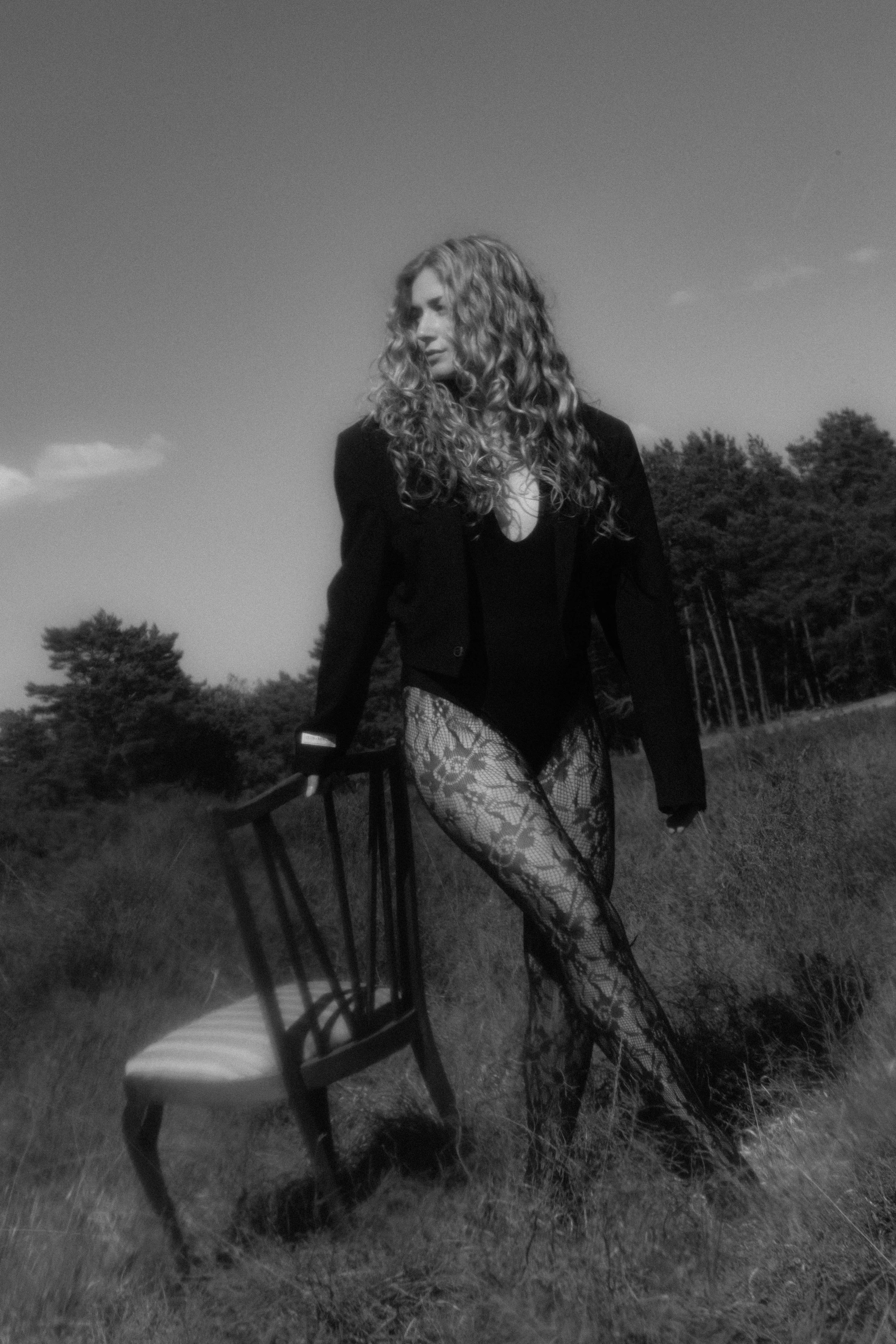 Een vrouw met lang, golvend haar staat in een grasveld met bomen op de achtergrond, in zwart-wwhite, gekleed in een blazer en lace panty, met een stoel die ze vasthoudt.