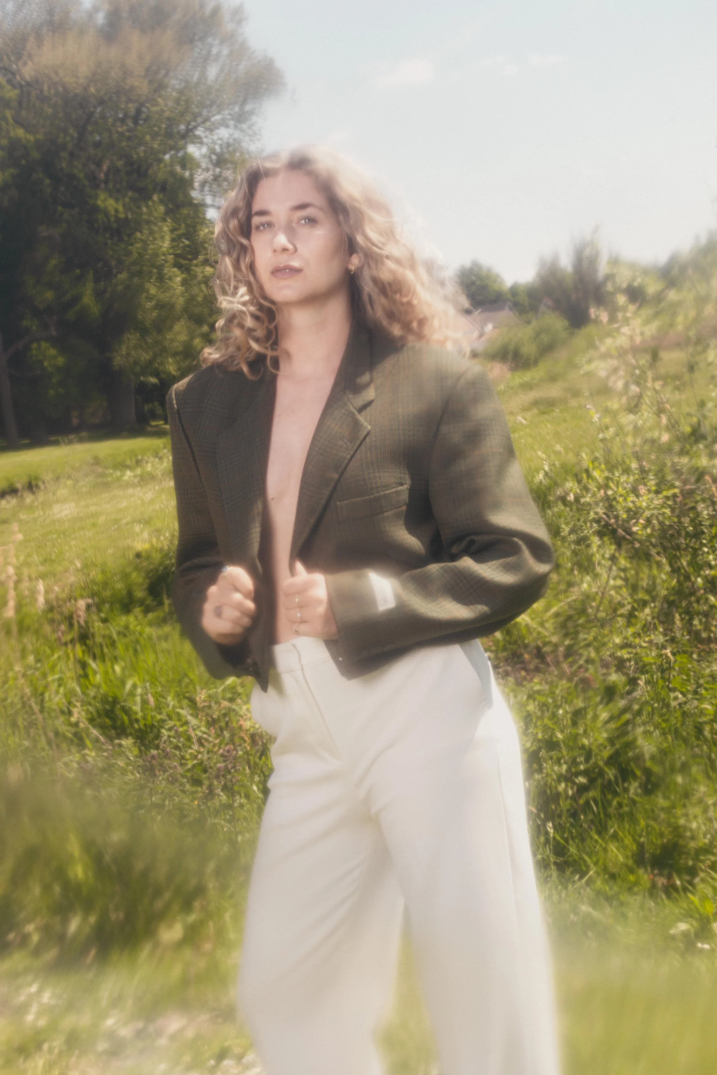 Een vrouw met golvend blond haar, gekleed in een oversized bruin colbert en witte broeken, staat op een grasveld met bomen op de achtergrond op een zonnige dag.