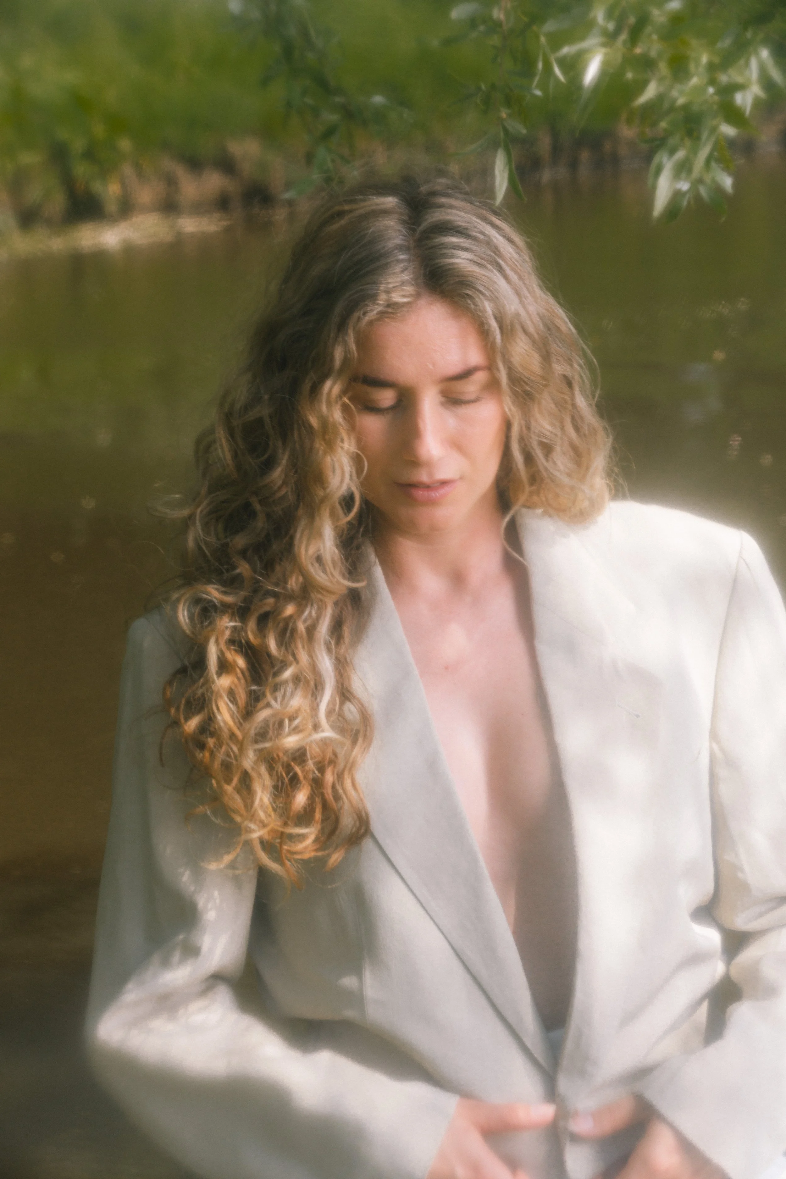 Vrouw met lang, gekruld haar, witte jas, dicht bij een rivier, omgeven door groene bladeren.
