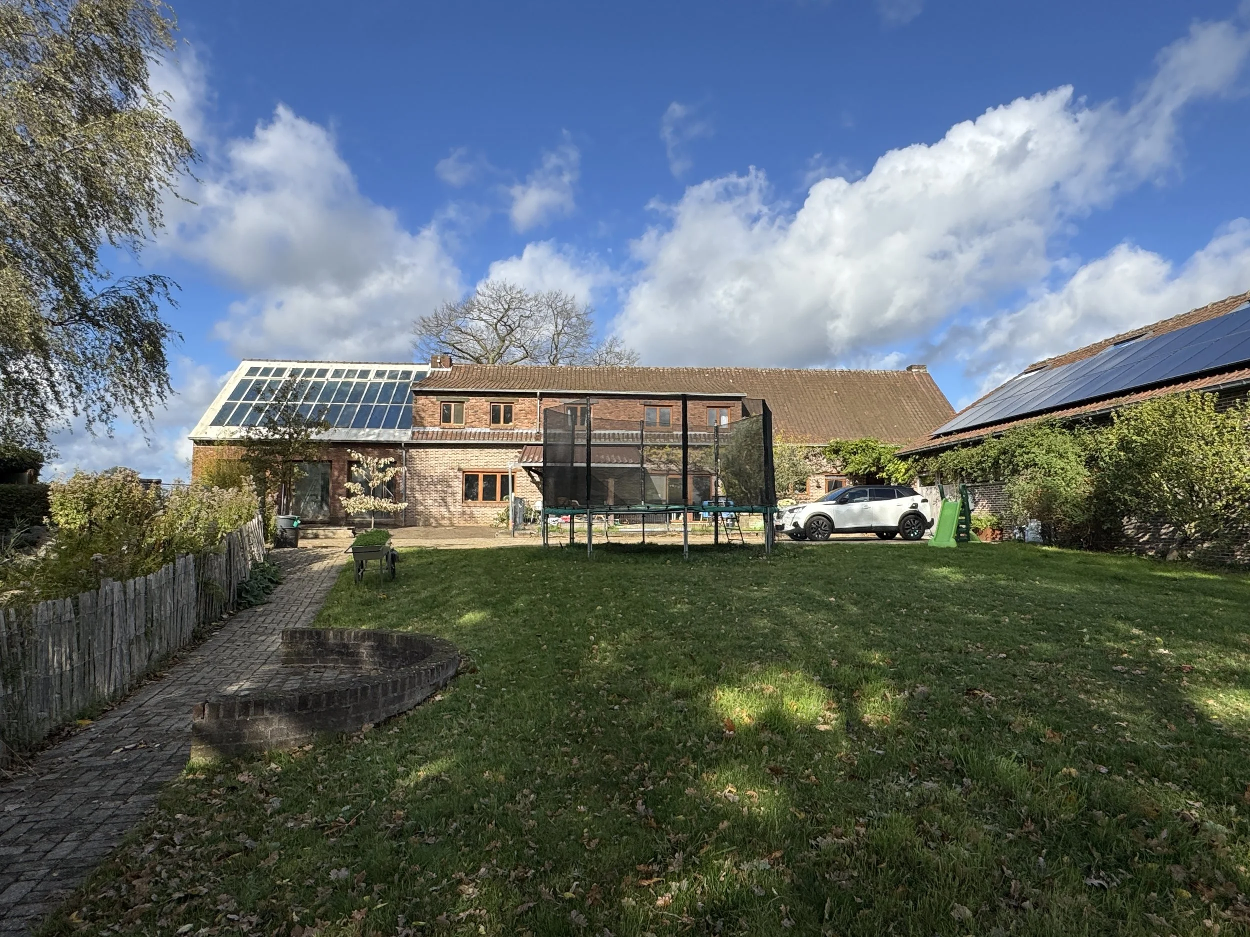 A backyard scene with a grassy lawn, a trampoline, a white car, a greenhouse with solar panels, and a house with a sloped roof. The sky is cloudy with patches of blue.
