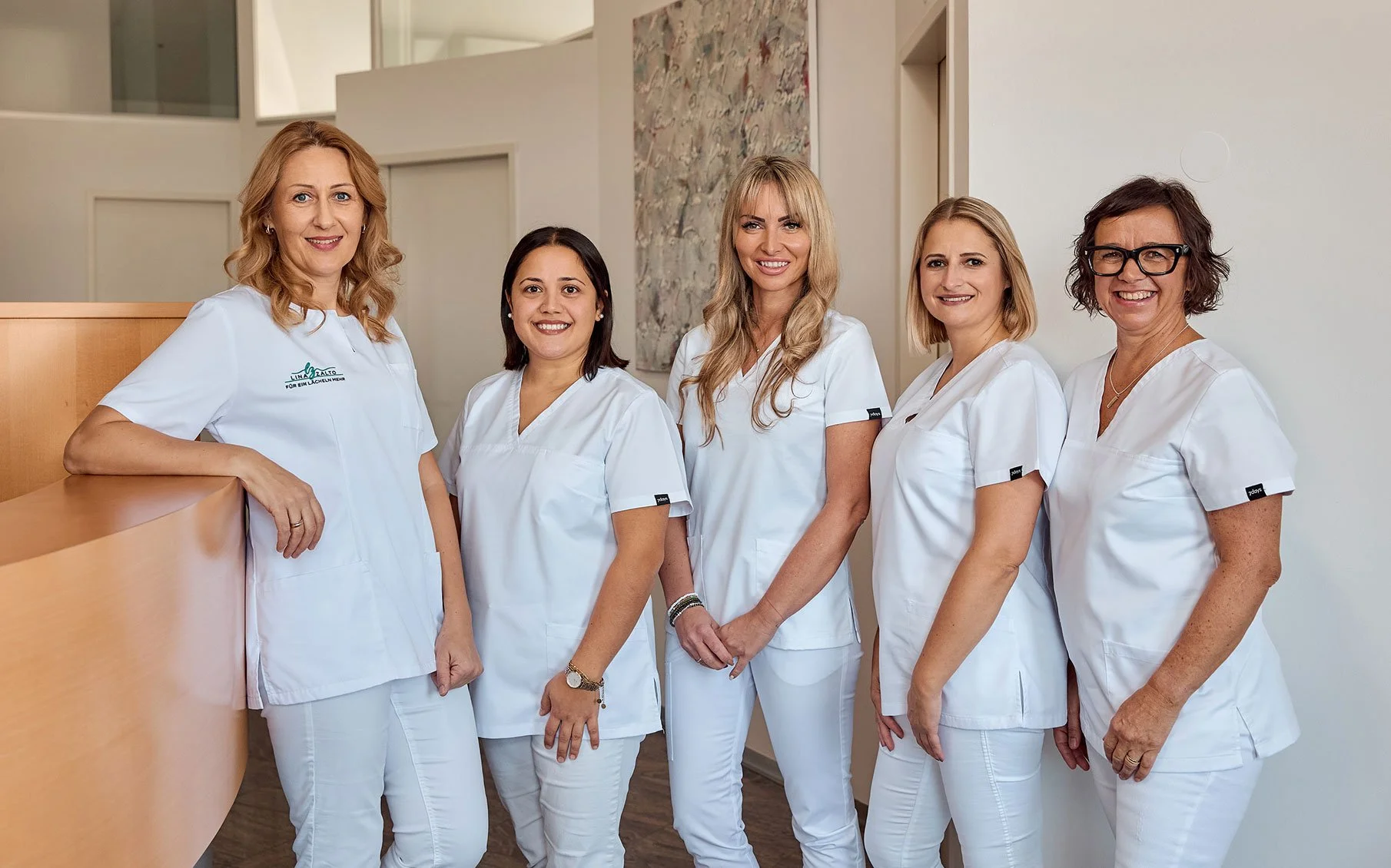 Fünf Frauen in weißen Pflegeuniformen stehen gemeinsam in einem medizinischen Empfangsbereich und lächeln in die Kamera.