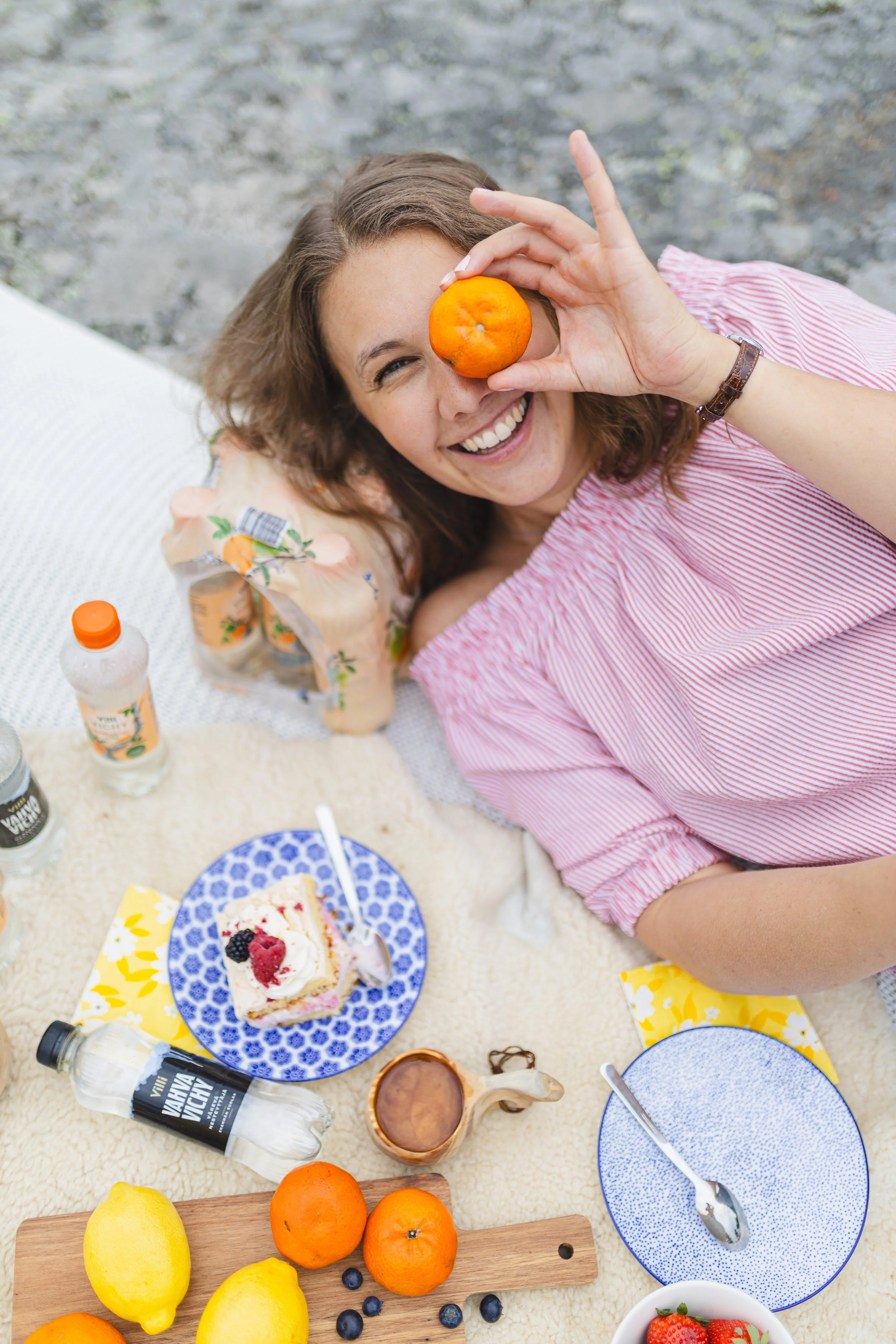 Nuori nainen hymyilee ja pitää appelsiinia silmänsä edessä pöydällä, juhlapiirakka ja hedelmiä pöydässä, mahdollisesti ulkona picnicissa.