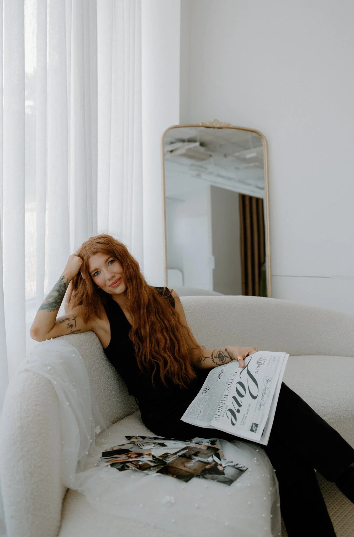 A women with curly red hair in all black outfit holding wedding photographs sitting on white couch with mirror behind her, tennessee wedding content creator