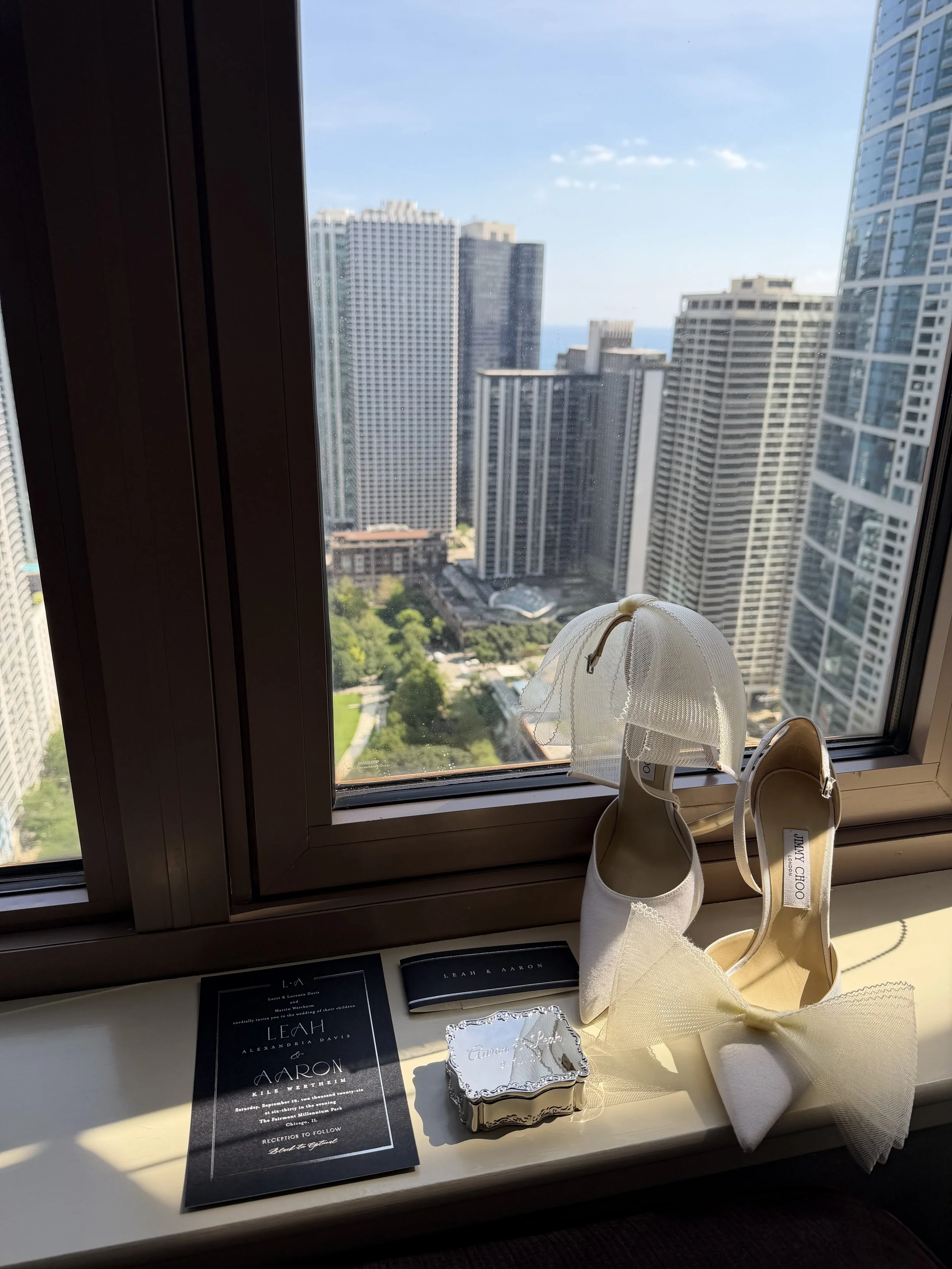 luxury wedding details including a black and white invitation, a silver engraved wedding ring box and white heels with bows sitting in front of a window of a pent house in Chicago wedding content creator