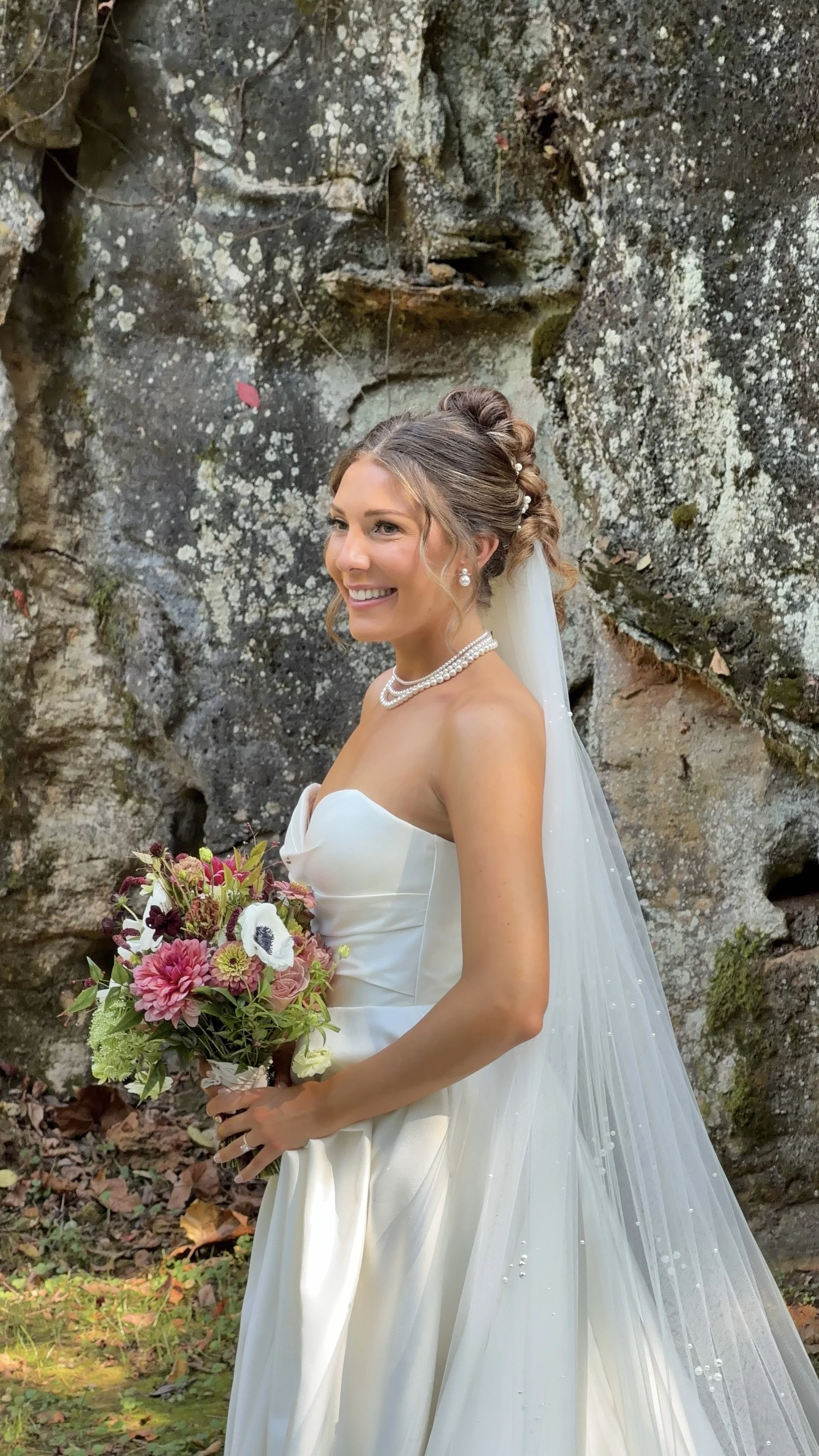 a young woman with blonde hair in a intricate bun in a wedding dress with a veil covered in pearls and pearl necklace and earrings. She is smiling at the camera and holding a bouquet of purple and pink flowers in front of a rock wall in a Quarry