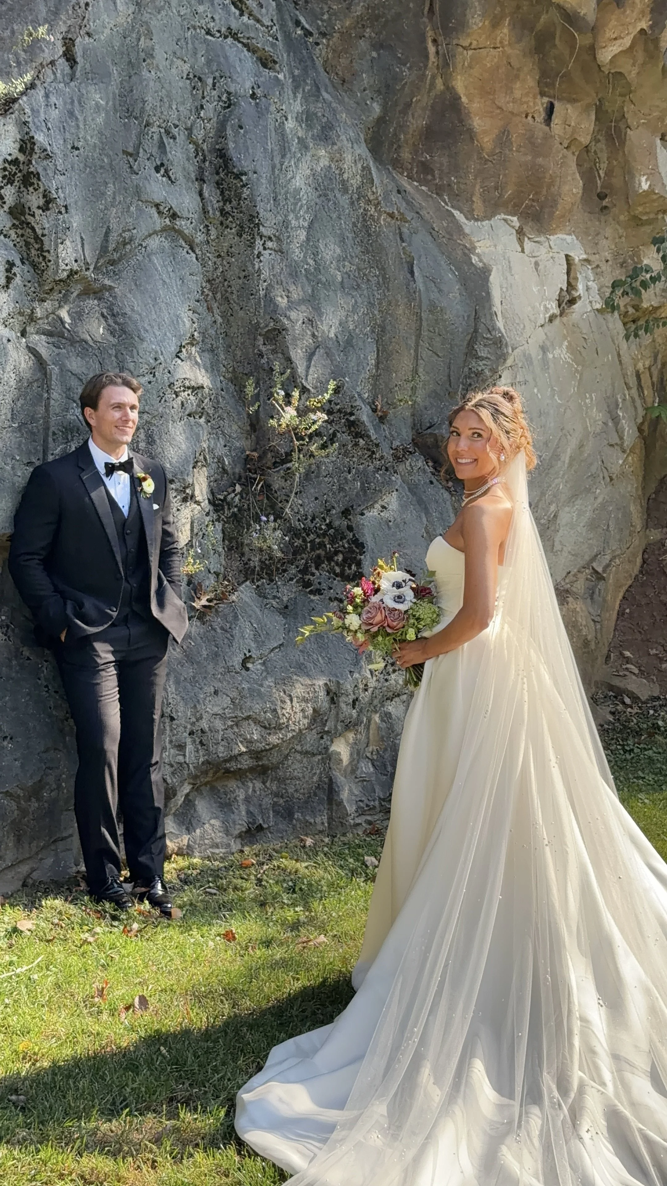 A young man in a black tux and a young women in a beautiful wedding dress with a pearl necklace and pearl earrings looking toward the camera and smiling, the background is a quarry rock wall
