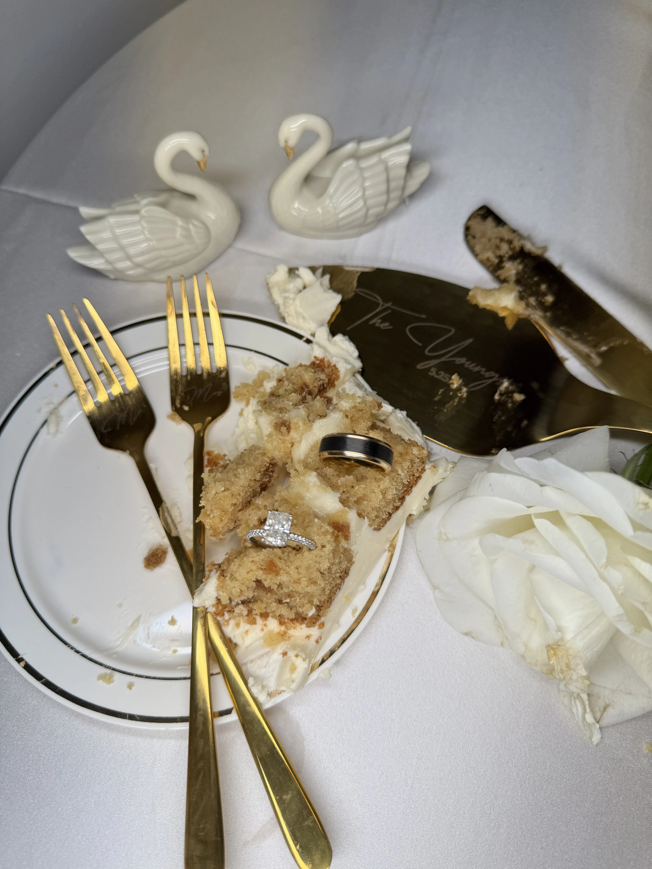 white cake with white frosting on a plate with gold forks and cake serving utensils with large diamond ring setting on the cake with white roses and two decorative swans luxury wedding content creator