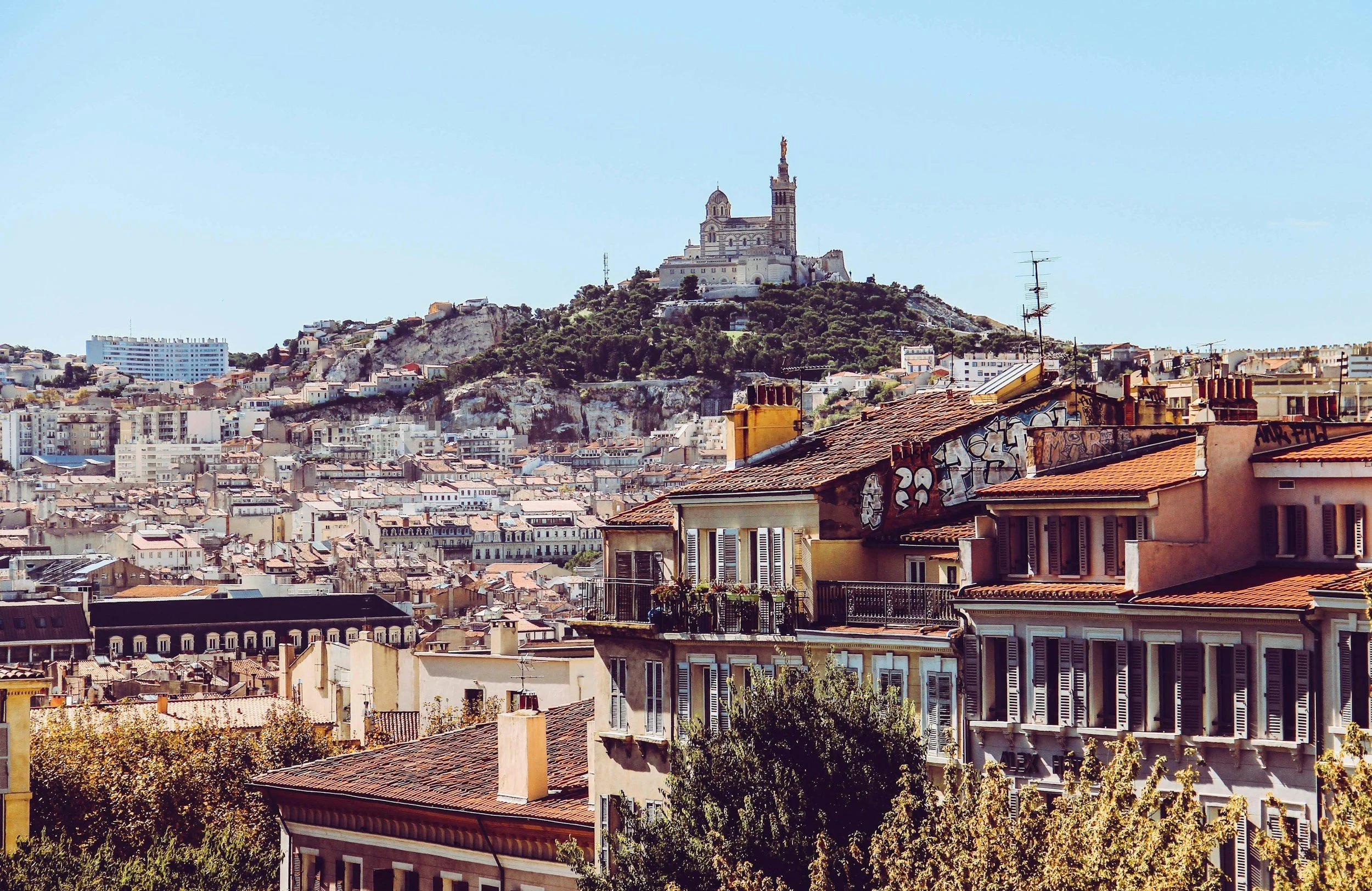 Vue sur les toits de Paris avec la basilique Notre-Dame de la Garde au sommet de la colline, en arrière-plan, sous un ciel bleu claire.