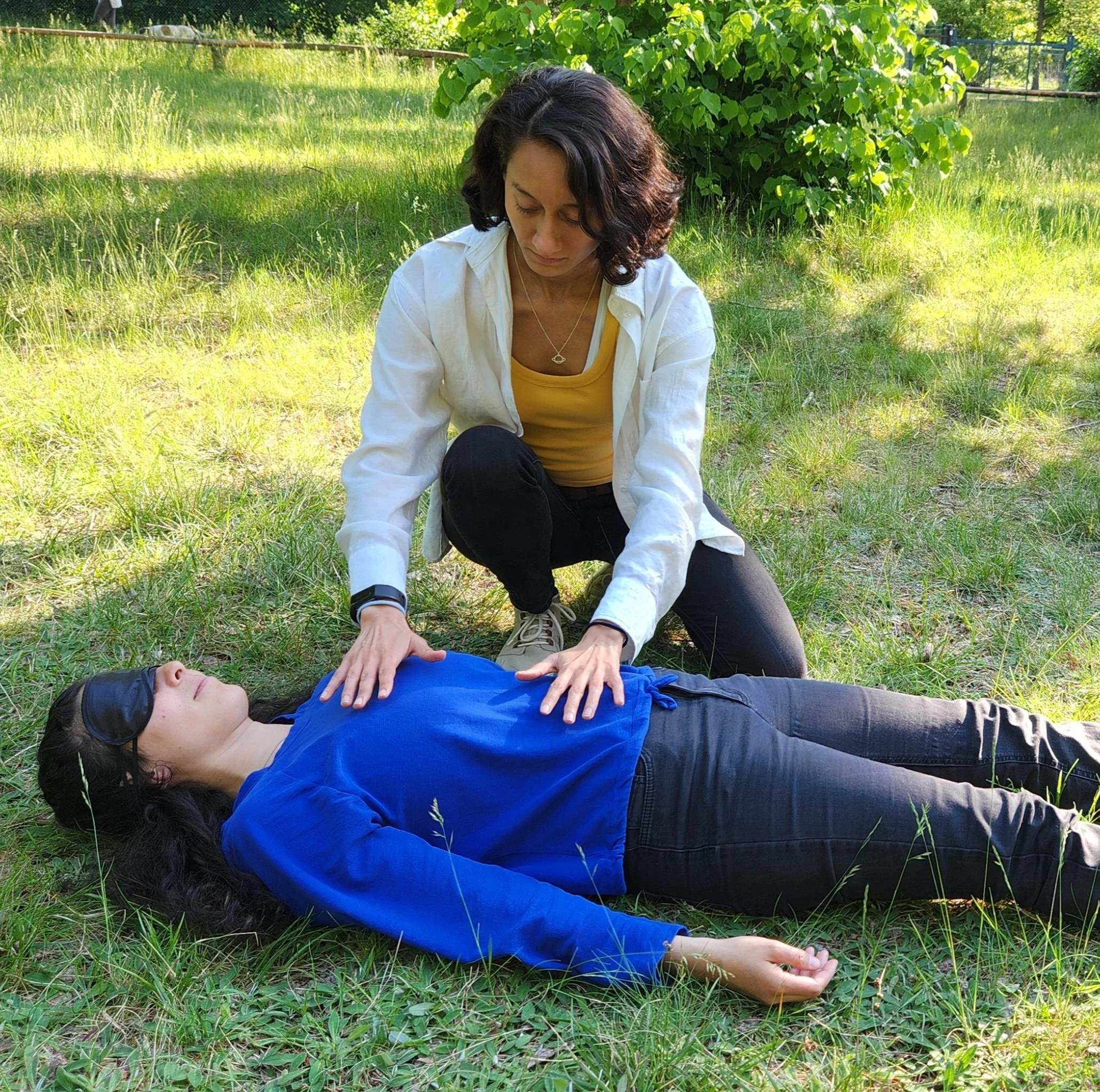 A woman performing CPR on another woman lying on the grass outdoors, with the woman on the ground wearing a blue shirt and a blindfold.