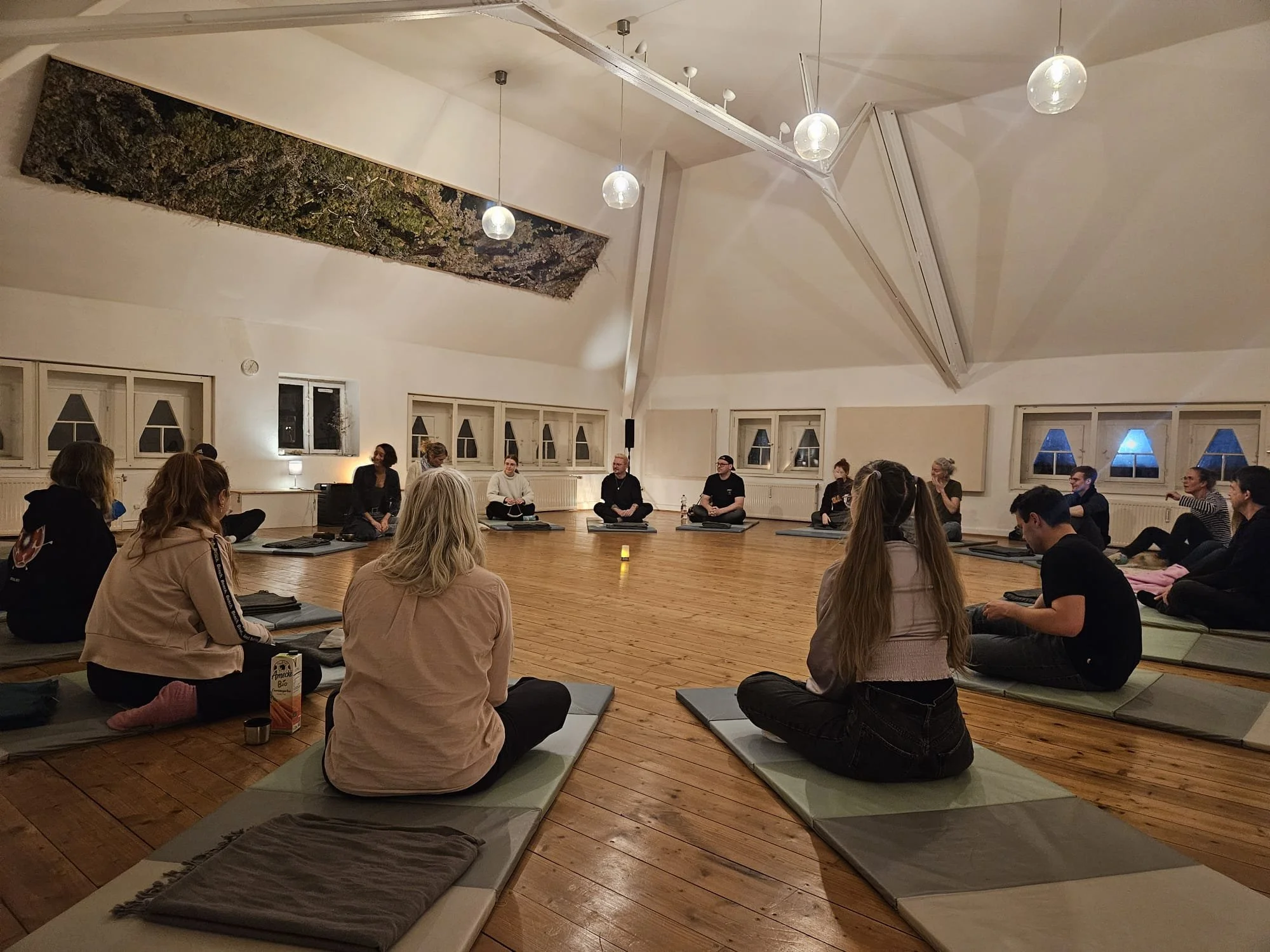 People sitting on mats in a circular arrangement in a spacious room with wooden floors, white walls, small windows, and a decorative ceiling feature, participating in a meditation or group activity at night.