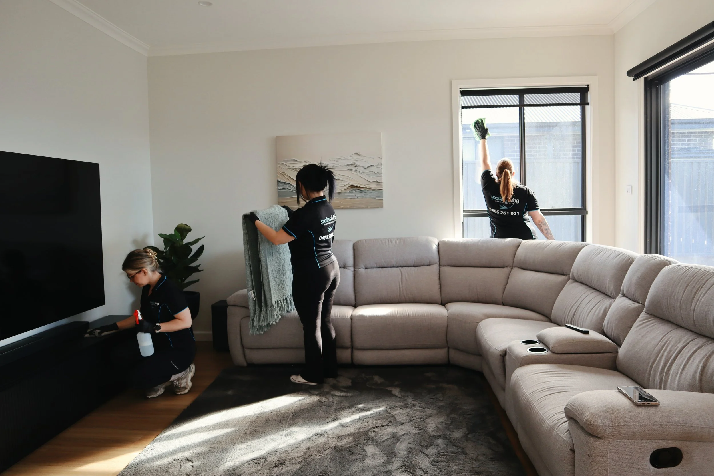 Three women in matching black cleaning uniforms working together to clean a living room with a large beige sectional sofa and a black entertainment center, with sunlight coming through the windows.