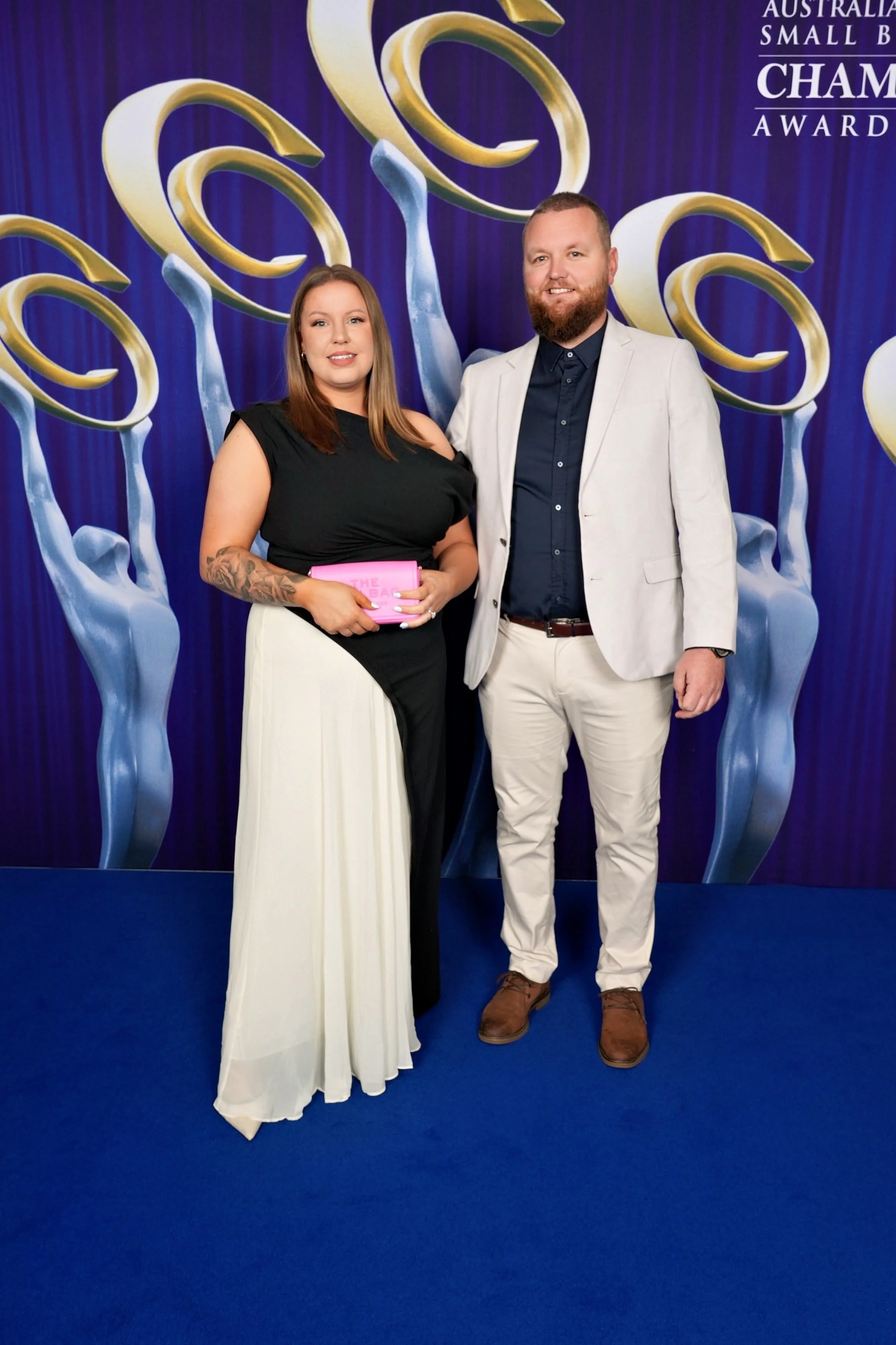 A man and woman standing indoors on a blue carpet in front of a decorative backdrop with gold and silver swirls. The woman has long hair, is dressed in a black top with one shoulder exposed and a long white skirt, and is holding a pink clutch. The man has a beard, is wearing a light-colored suit jacket over a dark shirt, and light-colored pants with brown shoes. They are smiling and posing for the photo at an awards event.