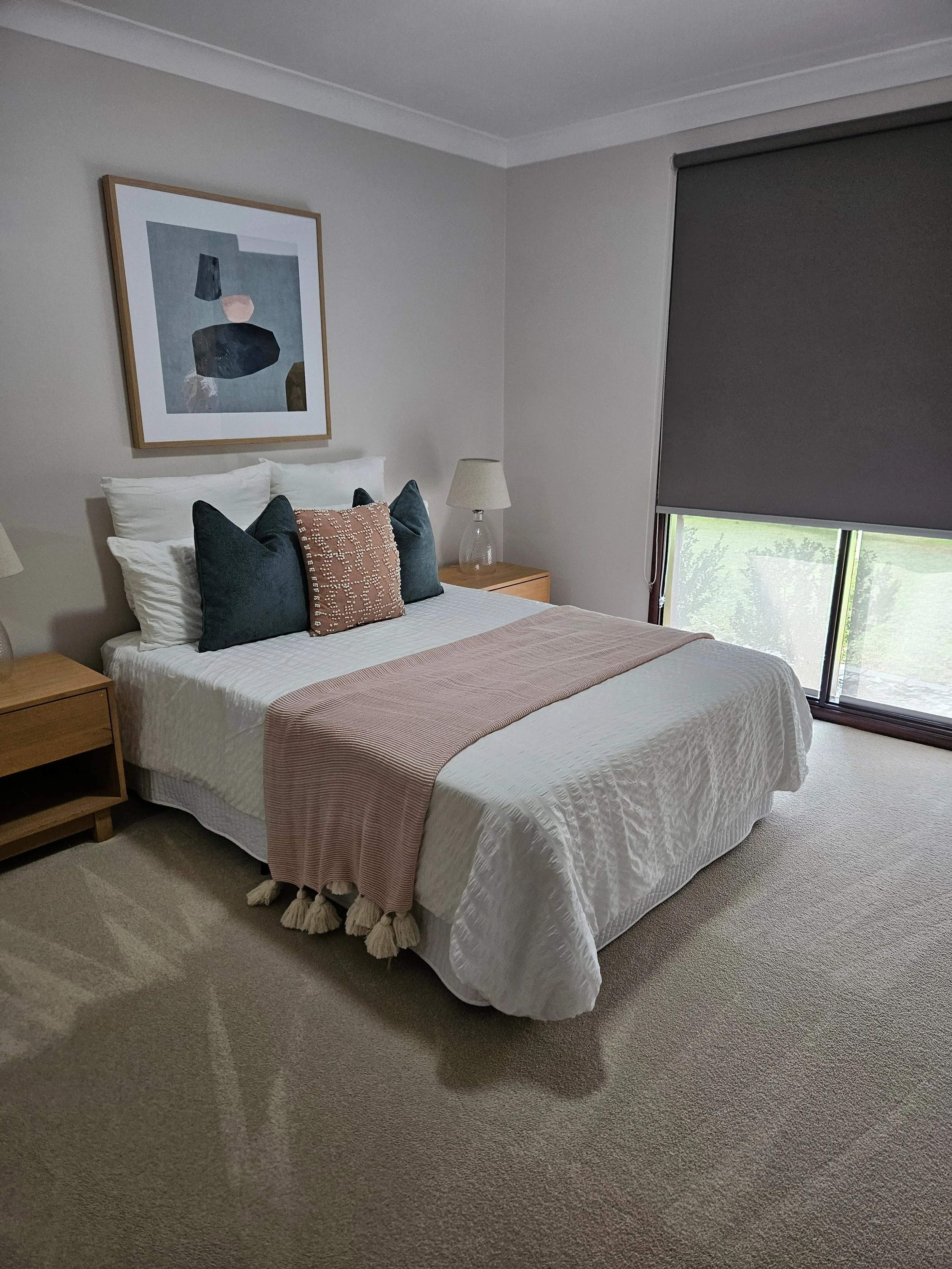 A neatly made bed with white bedding, a beige throw blanket, four pillows, and a wooden nightstand with a lamp on each side. There is an abstract artwork hanging above the bed and a window with a gray roller blind.