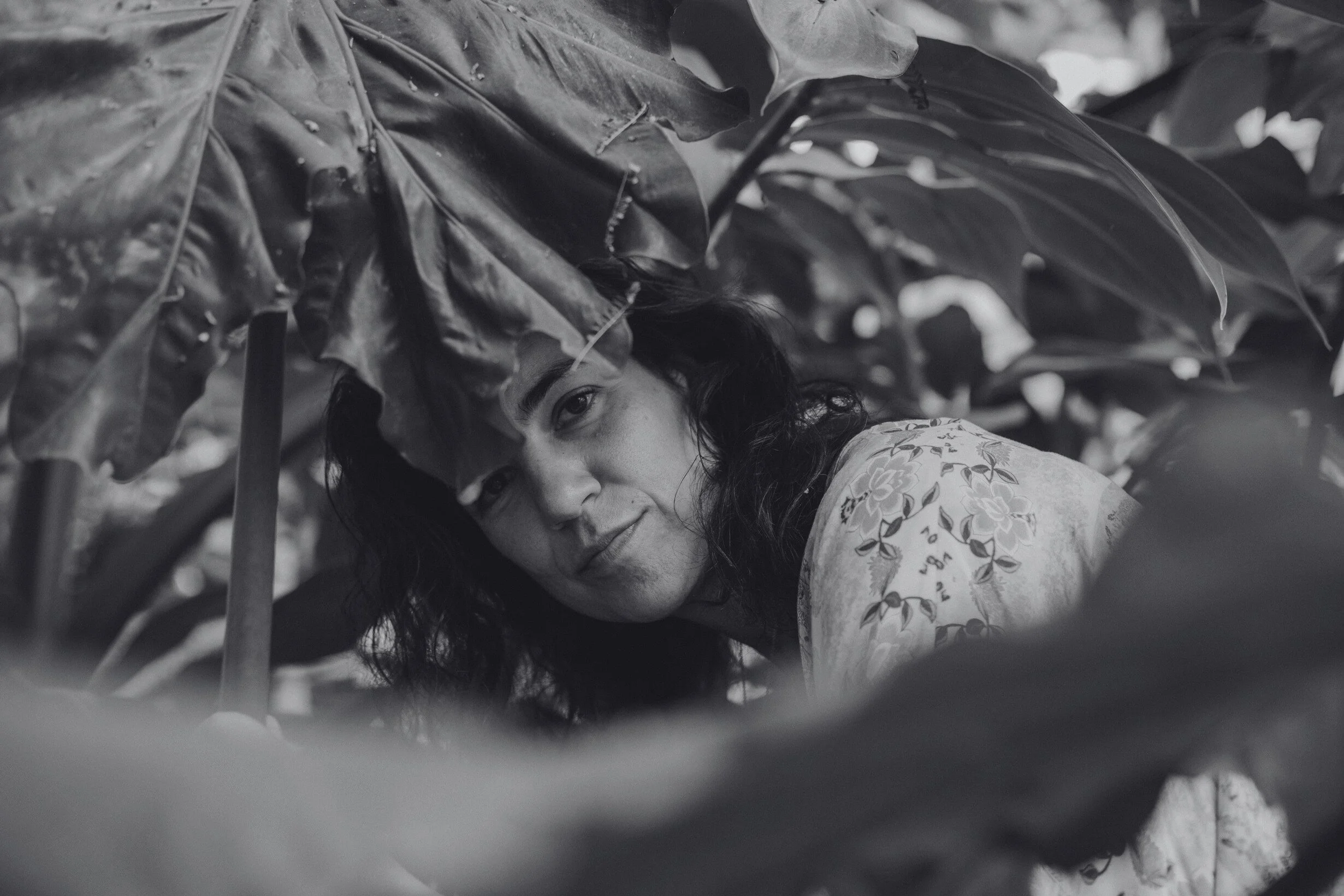 Mujer entre hojas de plantas grandes, mirando hacia la cámara en un entorno natural.