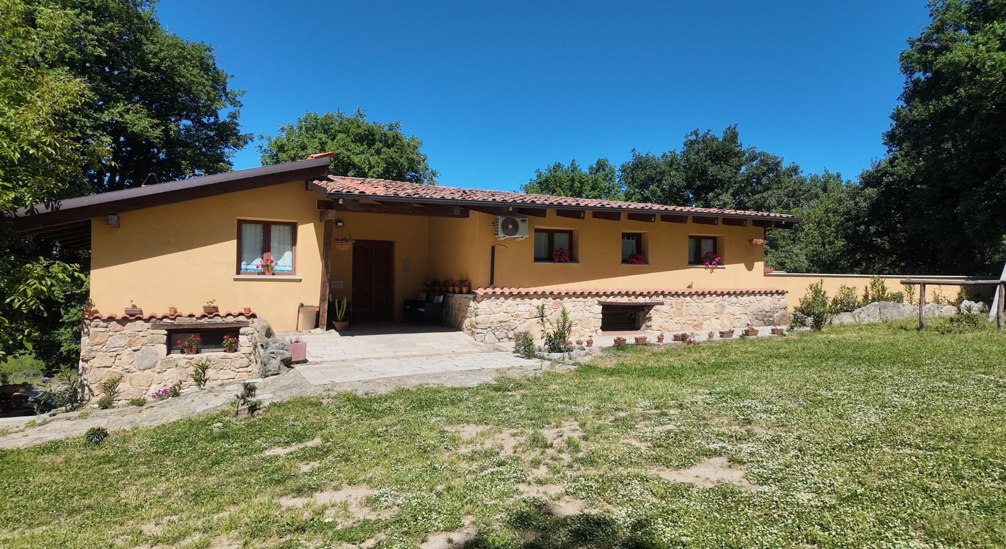 Casa in stile rustico con pareti gialle, tetto in tegole, finestre con fiori fioriti, terrazza con piante e ambiente verde circostante sotto un cielo blu.