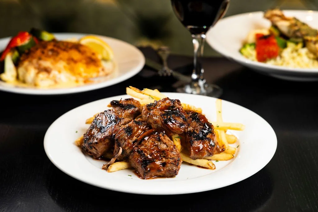 Plate with grilled ribs and French fries. In the background, plates with baked dish and a salad. Glass of red wine.
