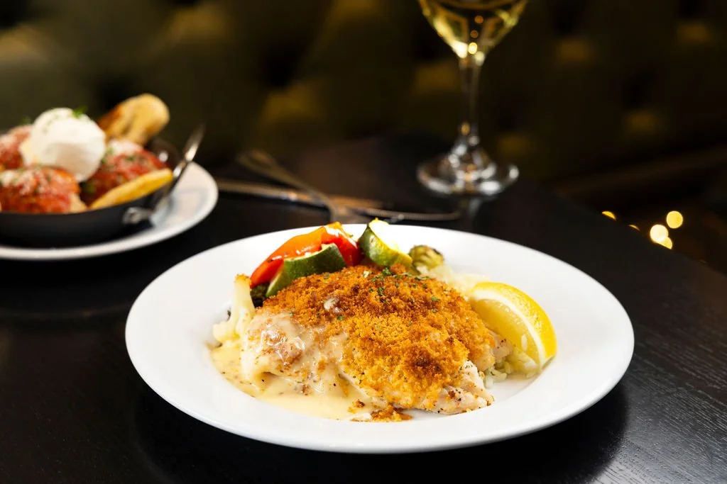 A plate of baked fish fillet topped with bread crumbs, served with lemon wedges and mixed vegetables on the side. In the background, a bowl of meatballs with sauce and a glass of white wine.