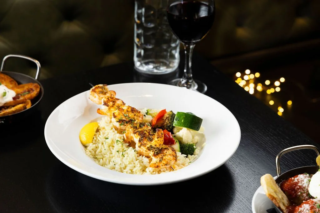Plate of grilled shrimp with rice and mixed vegetables, glass of red wine, and water on a dark table.
