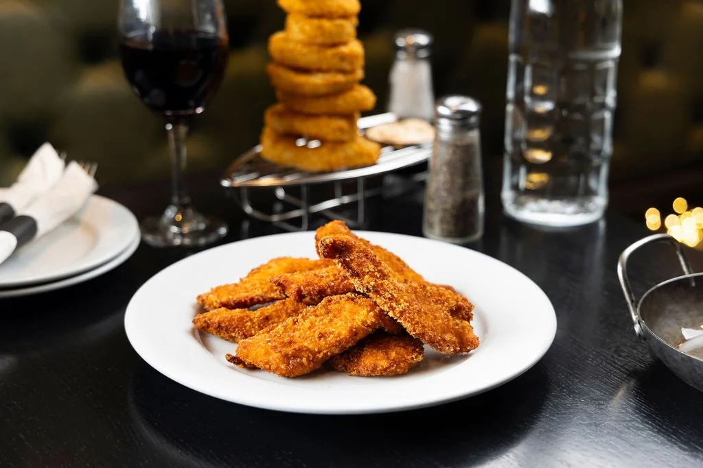 Plate of breaded fried fish fillets on a dark table, with a glass of red wine and a stack of onion rings in the background.