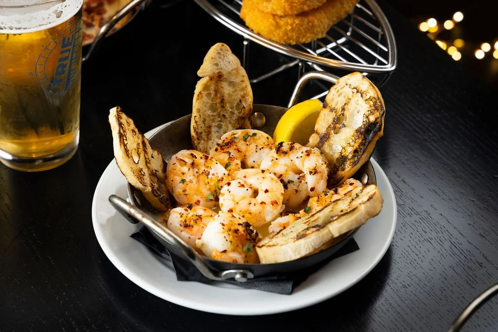 A bowl of grilled shrimp with toasted bread slices and a lemon wedge, served on a black table next to a pint of beer.