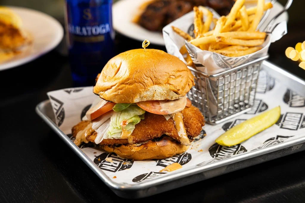 A breaded fish sandwich with lettuce, tomato, and sauce served with French fries and a pickle spear.